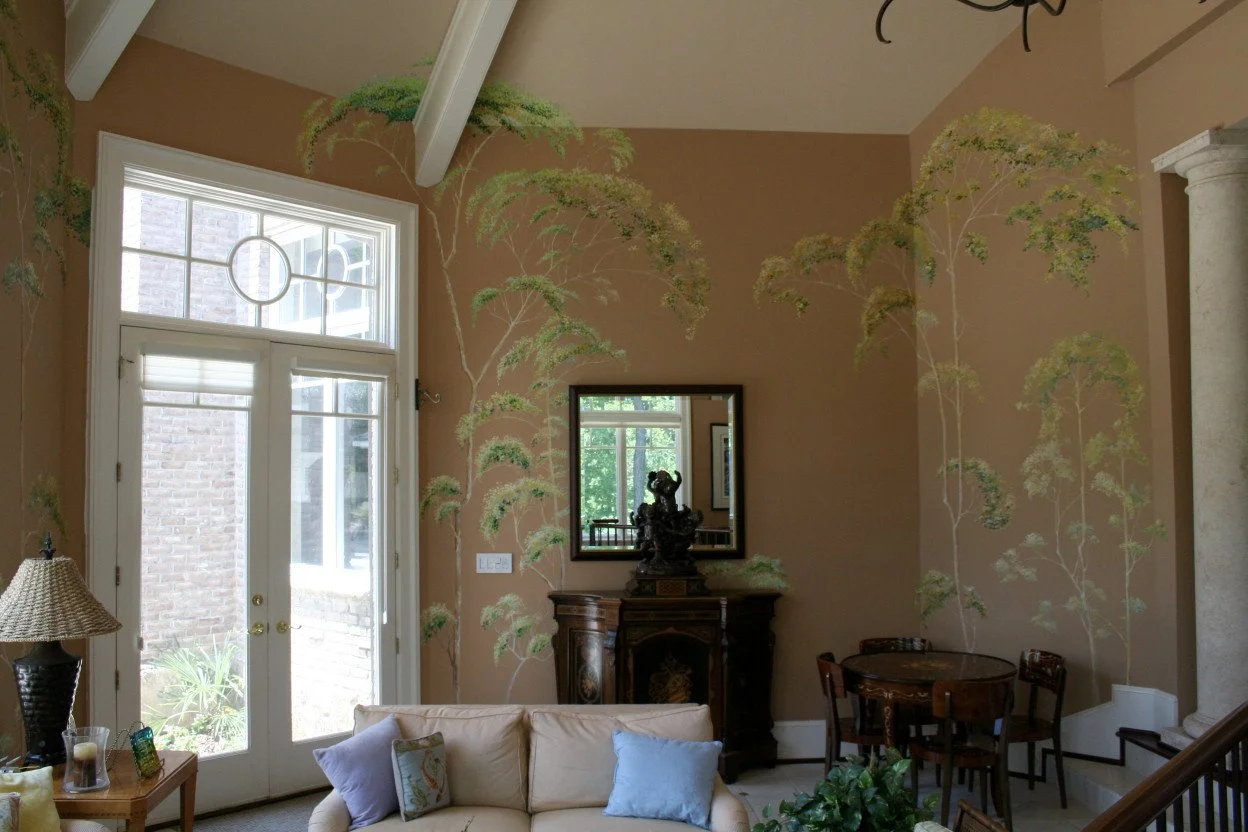 Living room with large window, beige walls with botanical mural, cream sofa with pillows, wooden furniture including side table with lamp, a small dining table with chairs, mirror on wall, decorative items, and a column.