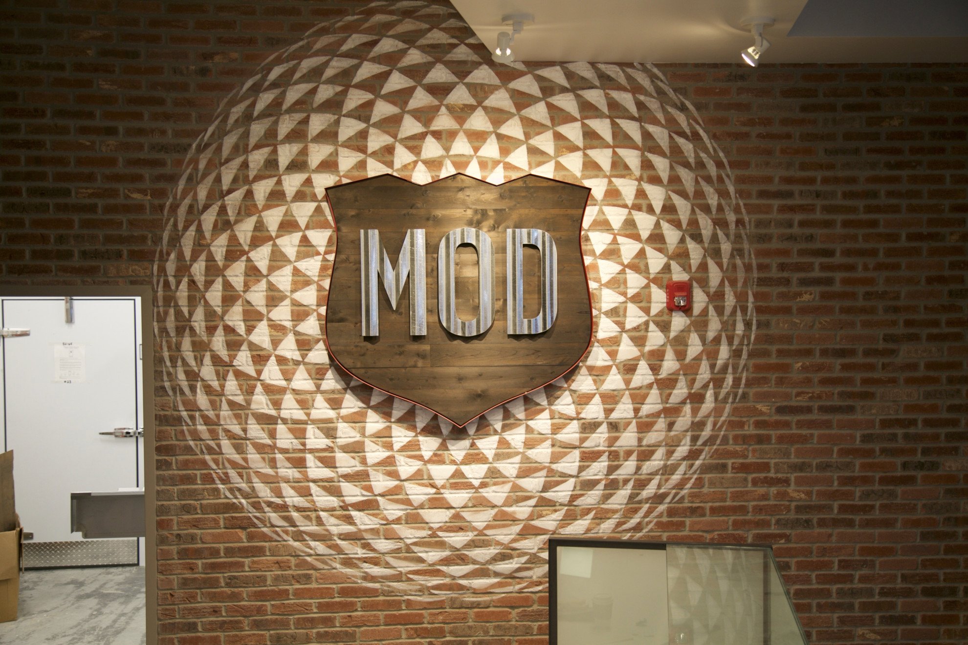 Decorative brick wall with a wooden shield-shaped sign displaying the word "MOD" and a large heart outline behind it, illuminated by ceiling spotlights.