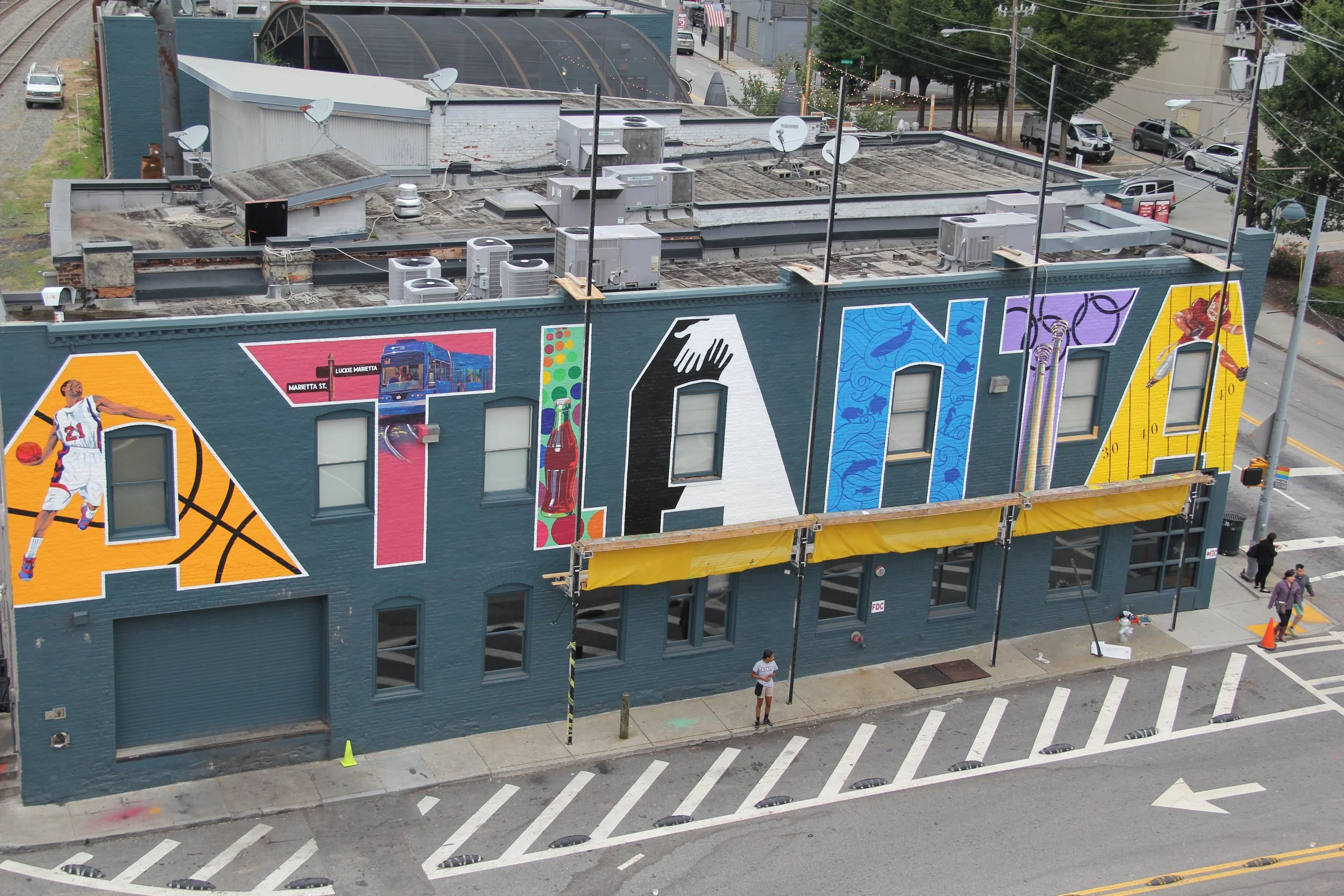A colorful mural painted on the side of a building in an urban area. The mural features images of a basketball player, a city bus, a wine bottle, water, and other artistic designs. The roof of the building has several air conditioning units and satellite dishes. There are people and cars visible on the street below.