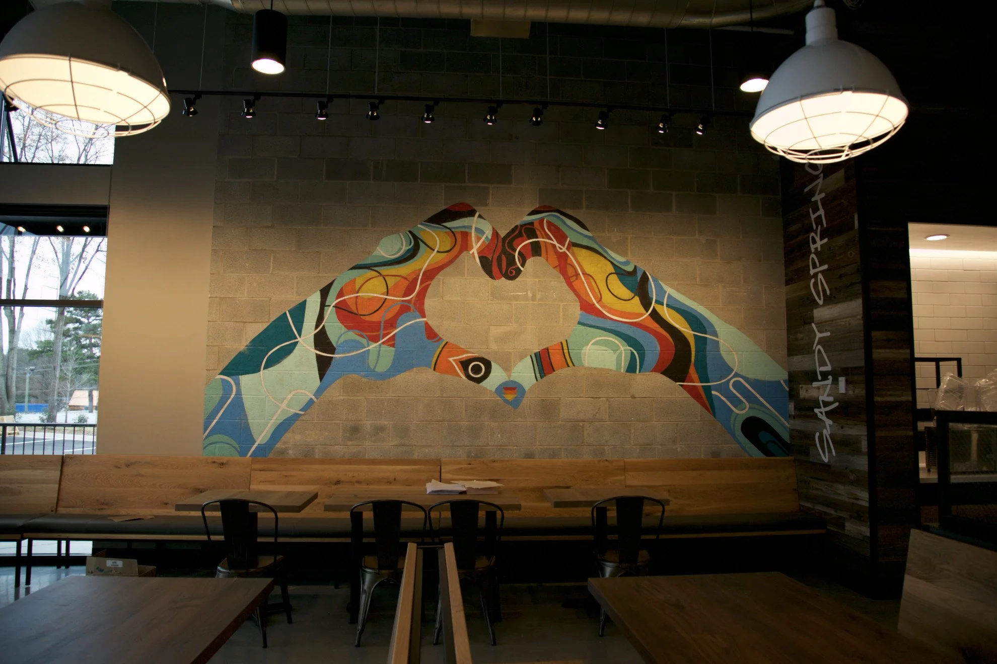 Colorful mural of two hands touching, forming a heart shape, on a brick wall in a modern cafe or restaurant with hanging lights and large windows.