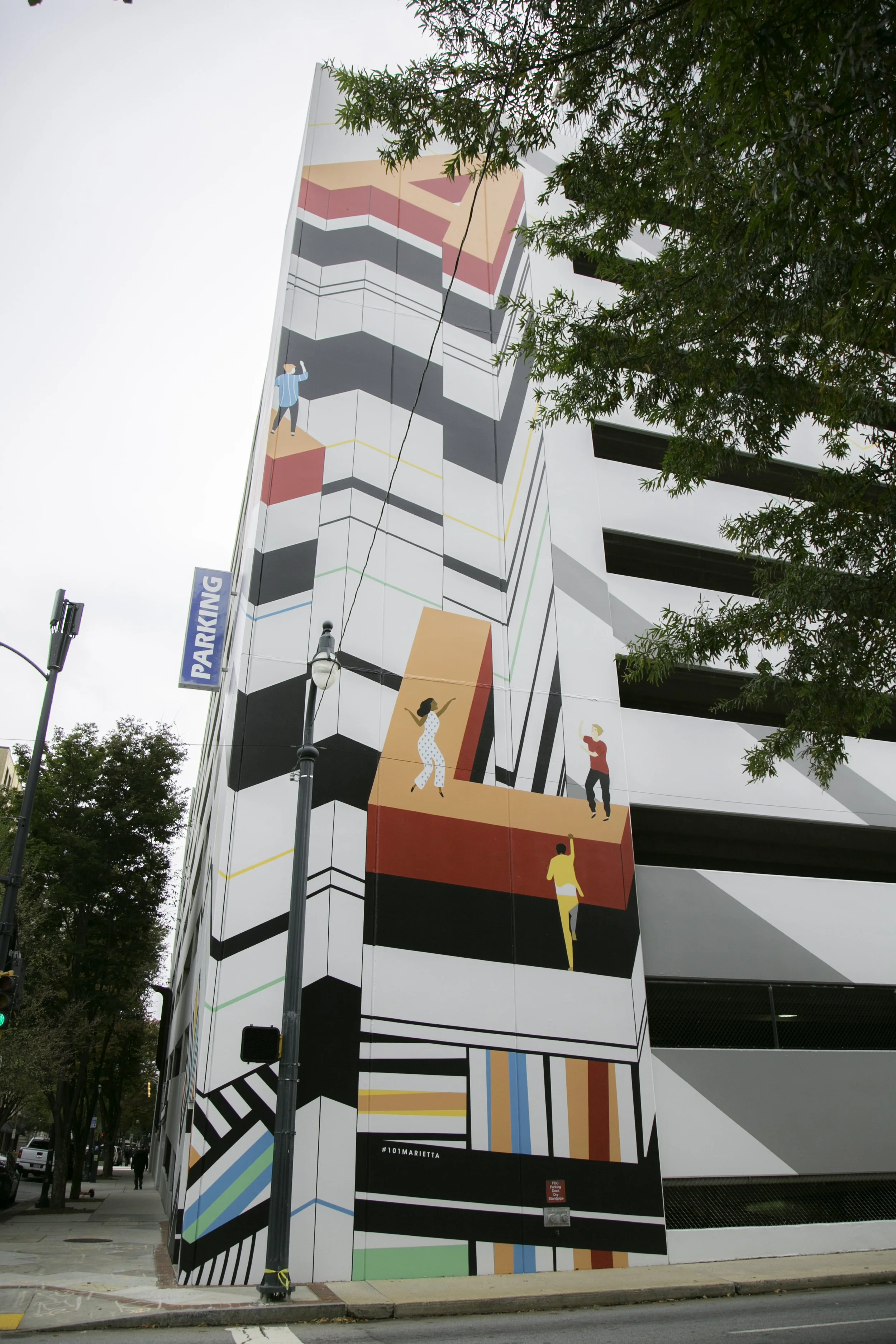Colorful mural on the side of a multi-story parking garage, featuring stylized illustrations of people, geometric patterns, and lines.