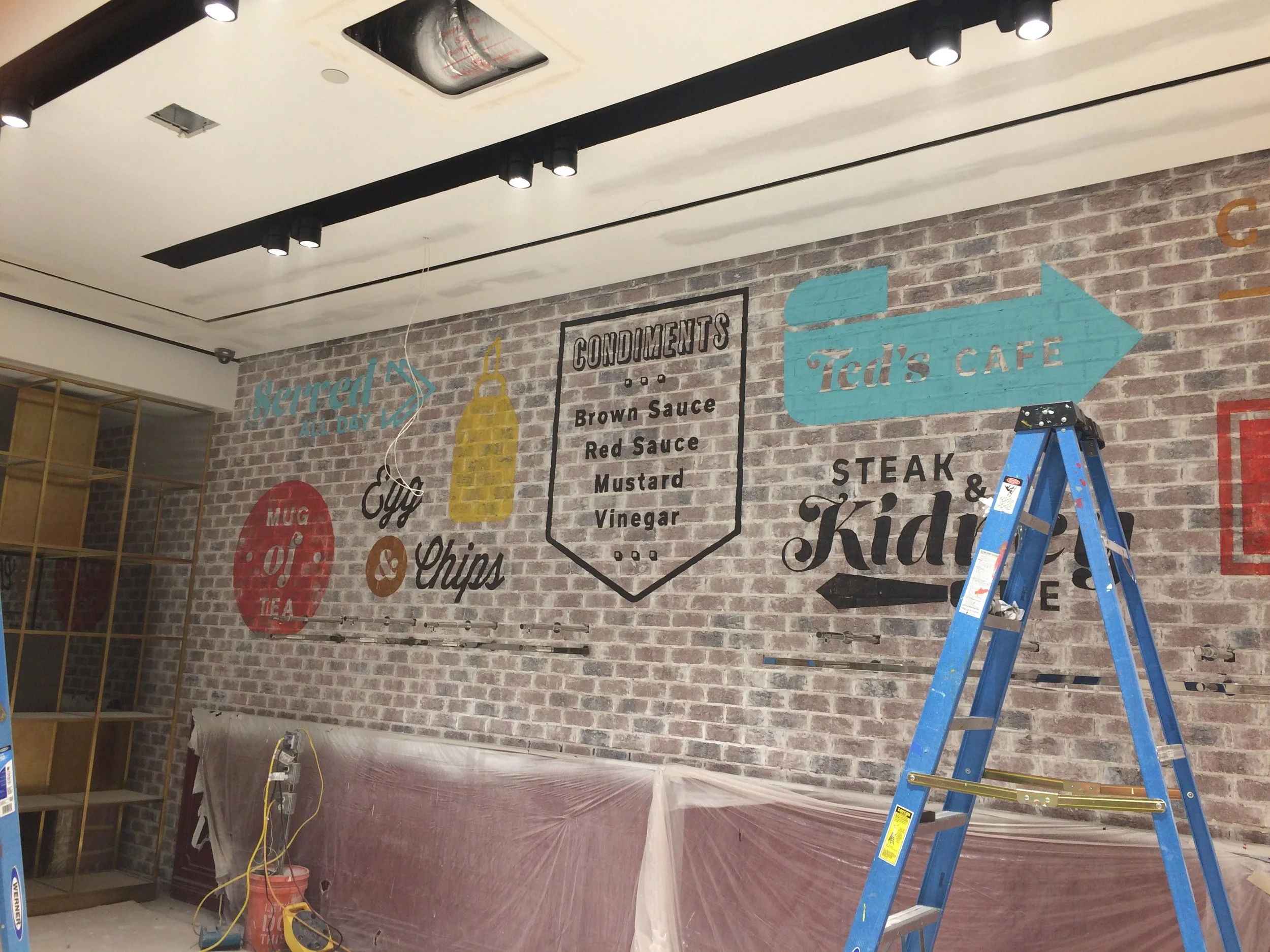 Interior of a restaurant under construction with a brick wall, black track lighting, a blue ladder, and a plastic-covered counter or bar area.