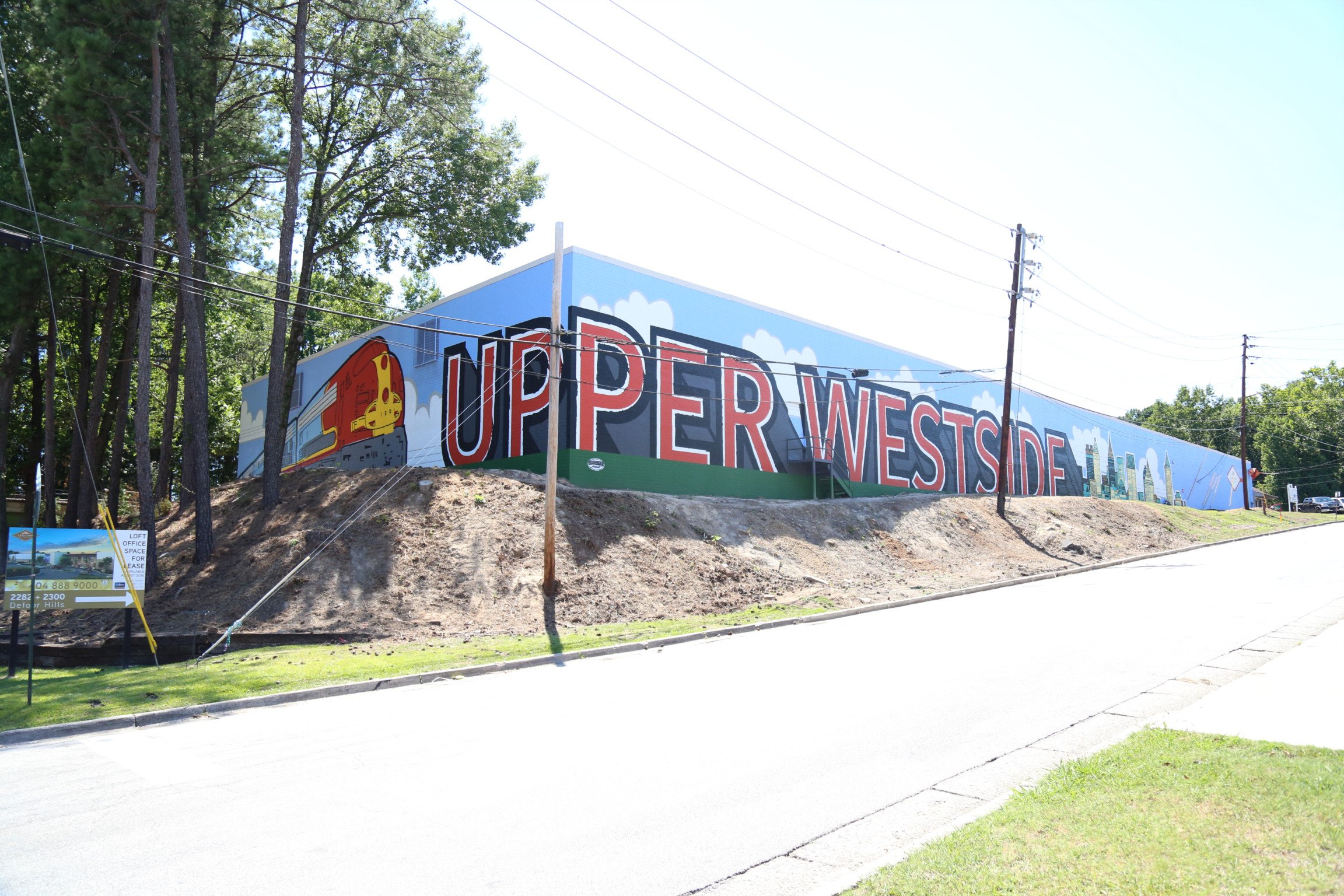 Large mural on a building wall with the text 'UPPER WESTSIDE' in bold letters. The mural includes a colorful illustration of a train engine and city skyline in the background. There are trees and power lines in front of the mural.