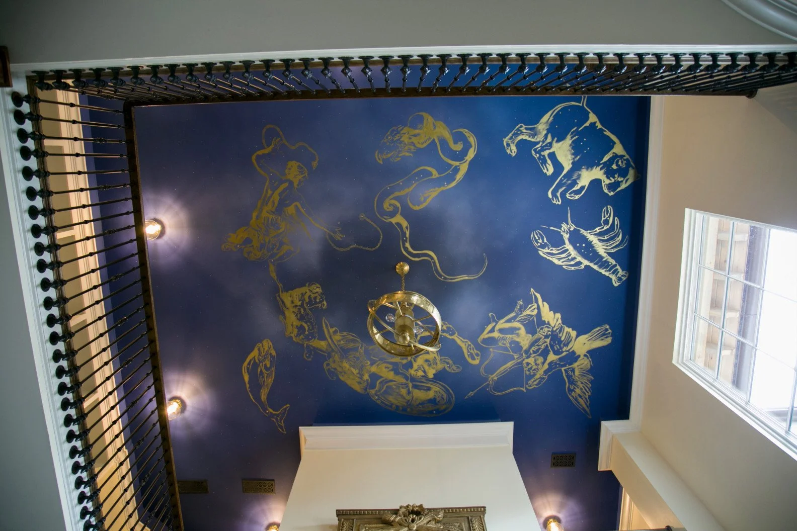 Ceiling painted with zodiac constellations in gold on a blue background, with a central brass chandelier, observed from below near a window.