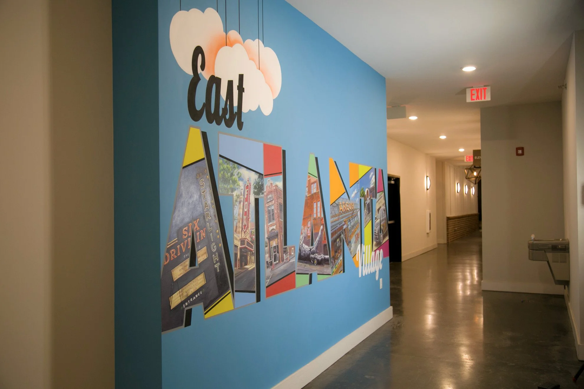 Interior of a building corridor with a blue wall featuring colorful posters and a sign that reads "Eat" with clouds above. The hallway has ceiling lights, an exit sign, and a bench.