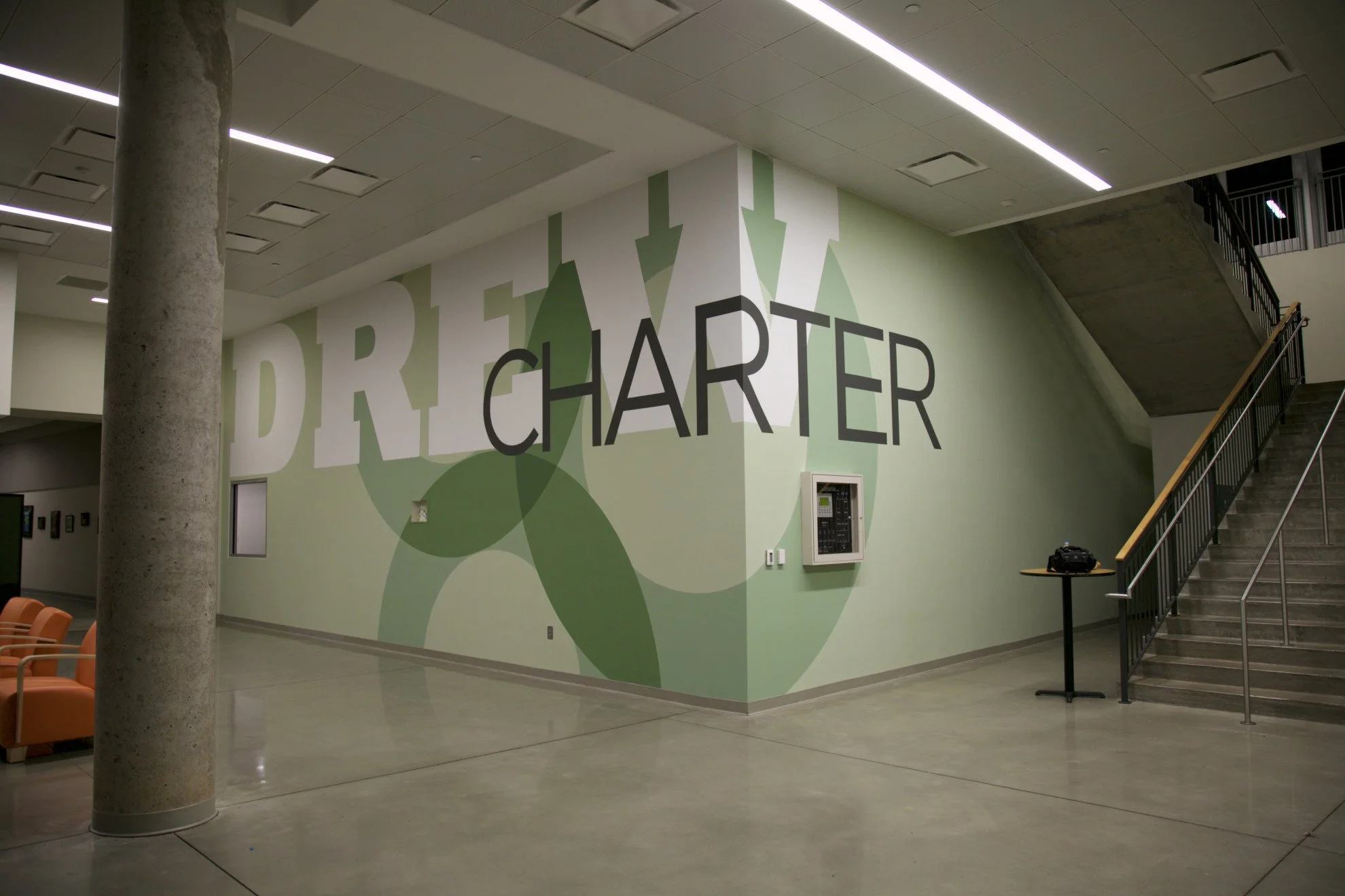 Indoor space with a large wall displaying the words 'DIRE CHARTER' and an abstract green graphic. There is a staircase on the right, an intercom or control panel on the wall, some chairs on the left, and a camera or equipment on a small table near the stairs. The ceiling has recessed lighting.