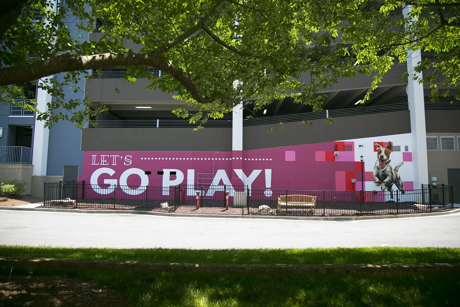 A colorful outdoor mural with the words 'Let's Go Play!' on a pink background, featuring a jumping dog and a lamp post, surrounded by a small black fence and greenery.