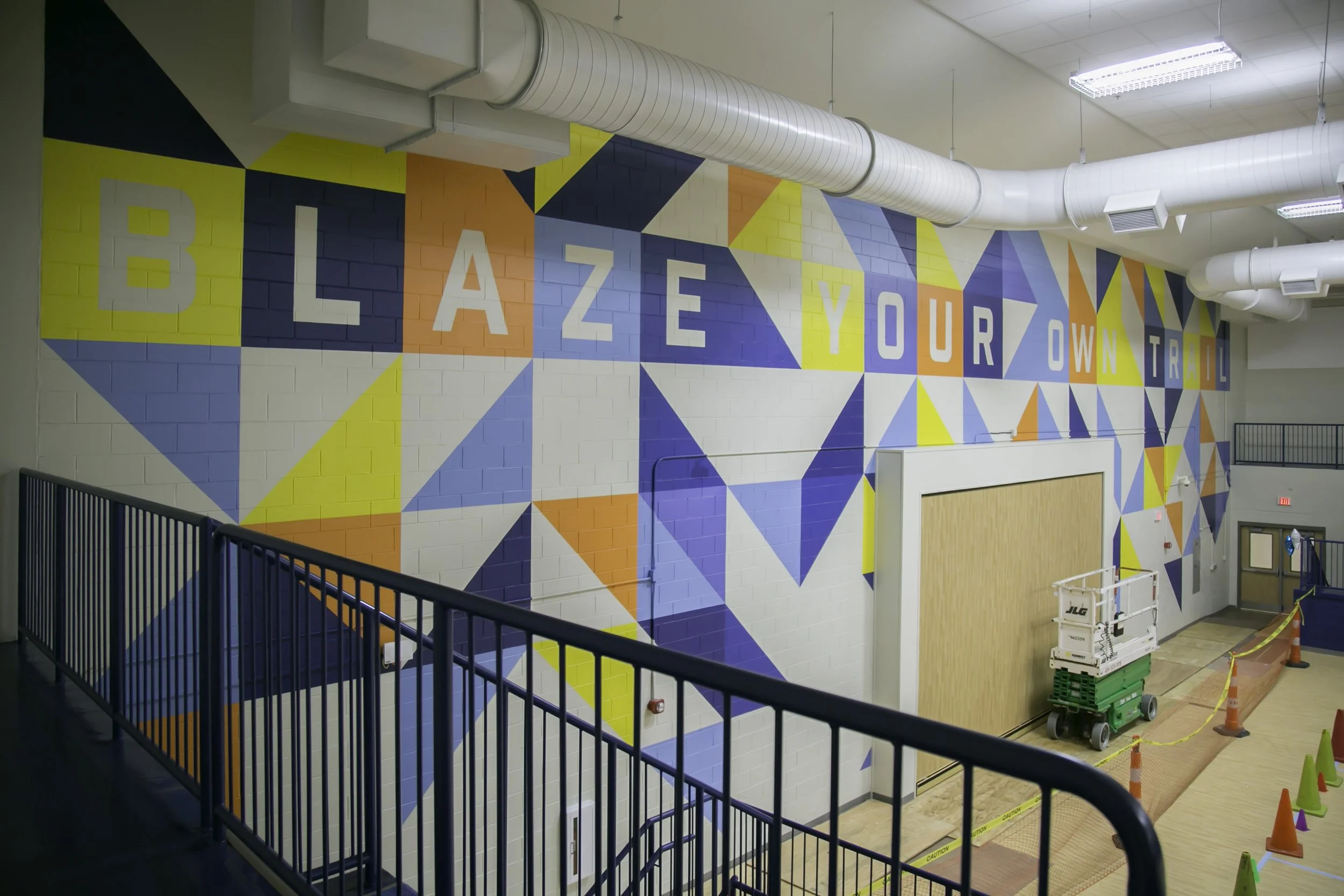 Colorful indoor mural with large letters spelling "BLAZE YOUR OWN TRAIL" on a brick wall, with ceiling ductwork and construction equipment visible.