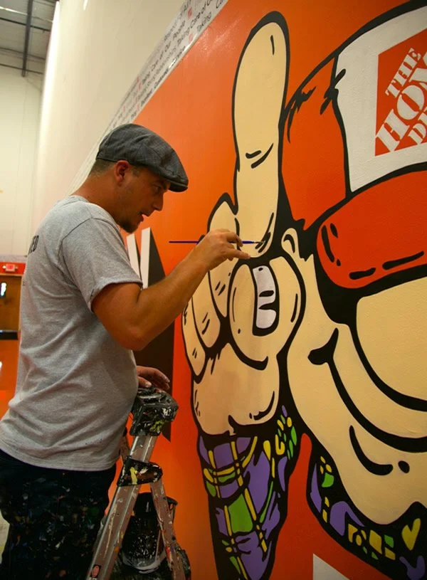 An artist painting a large mural on an orange wall that features a cartoon-style hand pointing with the index finger, holding a small box with the logo 'The Home Depot'. The artist is wearing a gray t-shirt, a gray cap, and has paint-splattered pants
