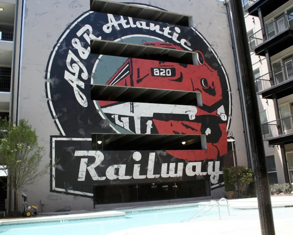 A large mural on a building featuring a red train engine with the numbers 820, surrounded by a circular design that reads "Atlatnic Speedway" and "Railway." Part of a swimming pool area is visible at the bottom.