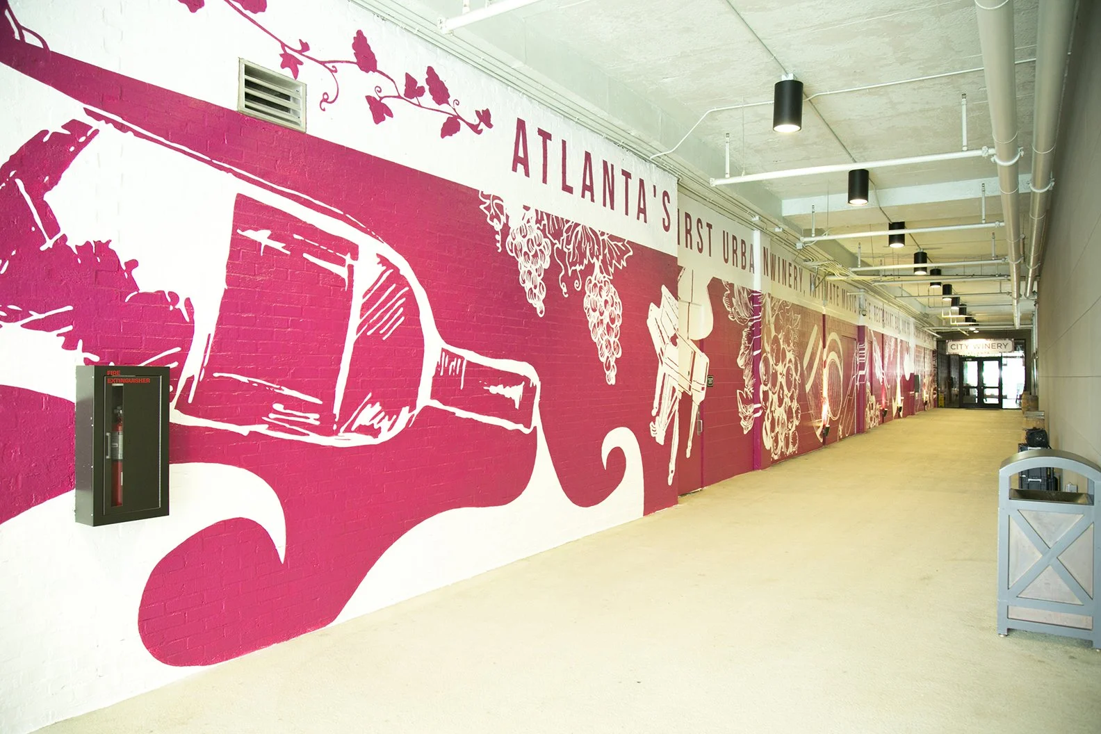 Indoor walkway with a mural on the wall featuring grapes, a wine bottle, and grapes, with the text "Atlanta's First Urban Winery". The ceiling shows exposed pipes and lighting fixtures, with a fire extinguisher box on the wall and a trash bin at the end of the walkway.