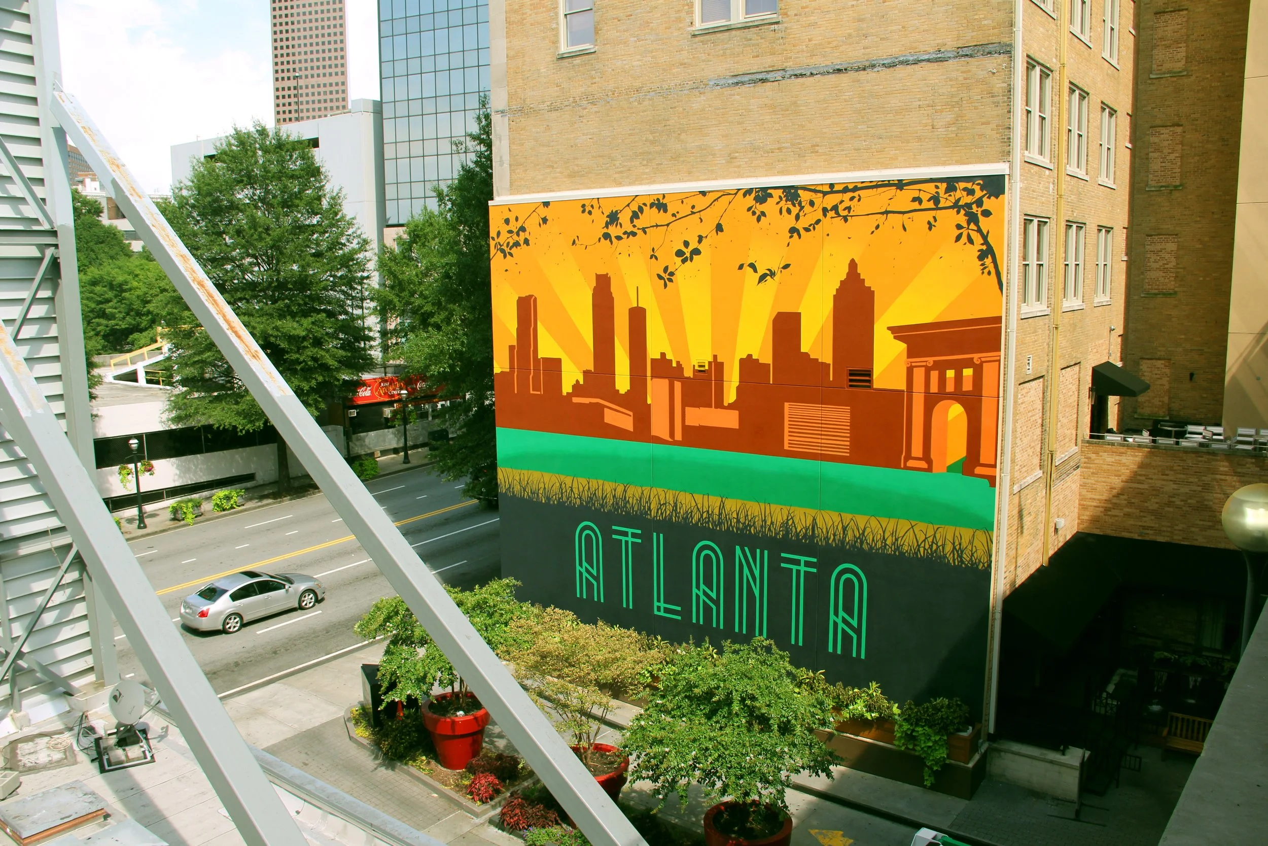 A city street with a large colorful mural on a brick building that depicts a city skyline with orange and yellow rays in the background, and includes the words 'ATLANTA' in neon-style lettering.