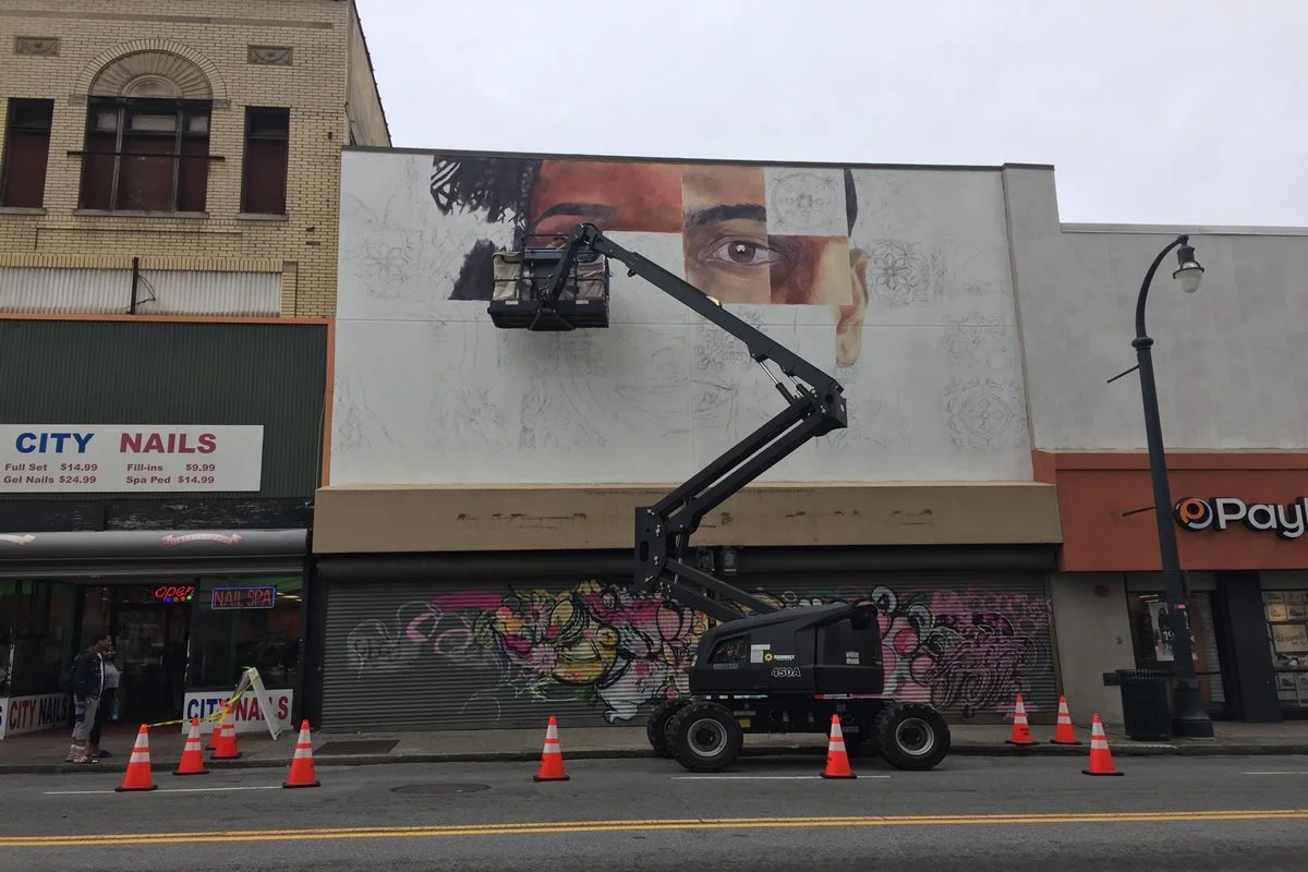 A worker in a lift is painting a large mural of a person's face on the side of a building. The face is partially completed, showing detailed eyes and hair, with sketches and outlines of the rest of the face remaining.