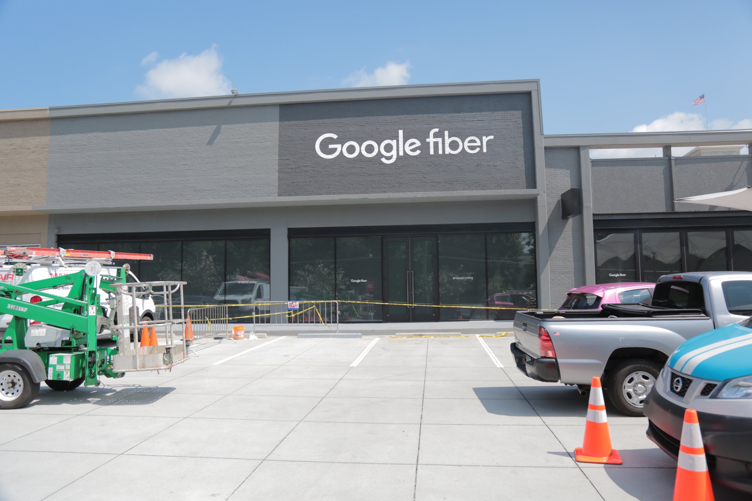 Google Fiber storefront under construction with construction vehicles, orange safety cones, and parked cars in the parking lot.