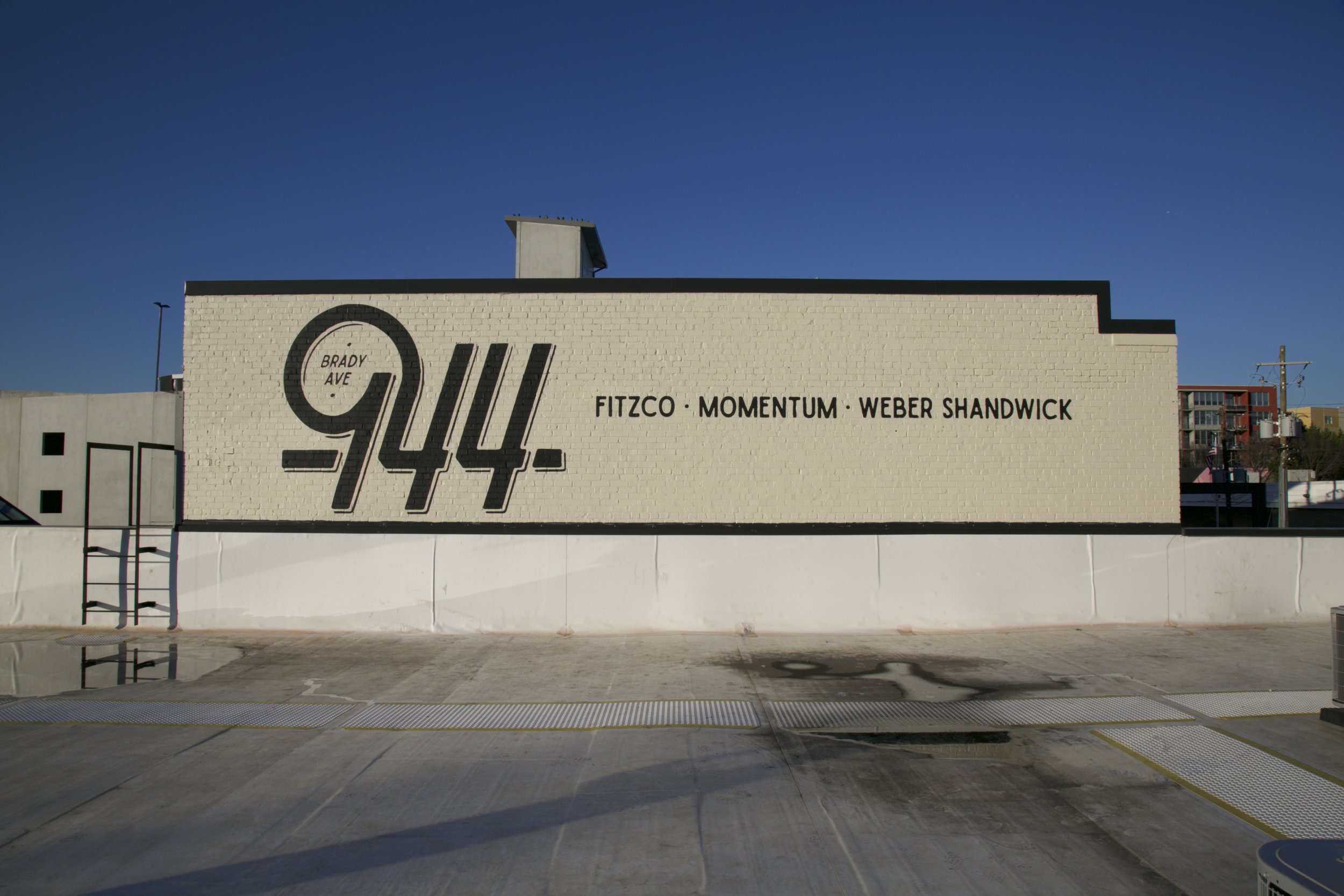 A building with a large painted mural featuring the number 944, and the names Brady Ave, Fitzco, Momentum, Weber Shandwick on the wall, with a clear blue sky and some distant buildings.