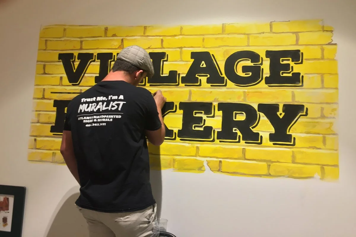 Person painting a mural of yellow brick wall with black text that reads 'VILLAGE LIBRARY'.
