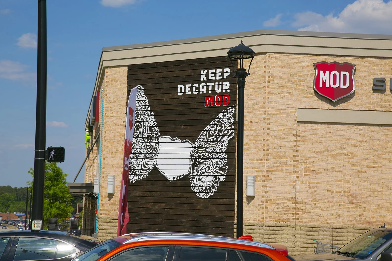 The exterior of a shopping center with a sign that says 'Keep Decatur MOD' and features a large graphic of a face with wings. There is a MOD shield logo on the building, a pedestrian crossing sign, and cars parked in the foreground.