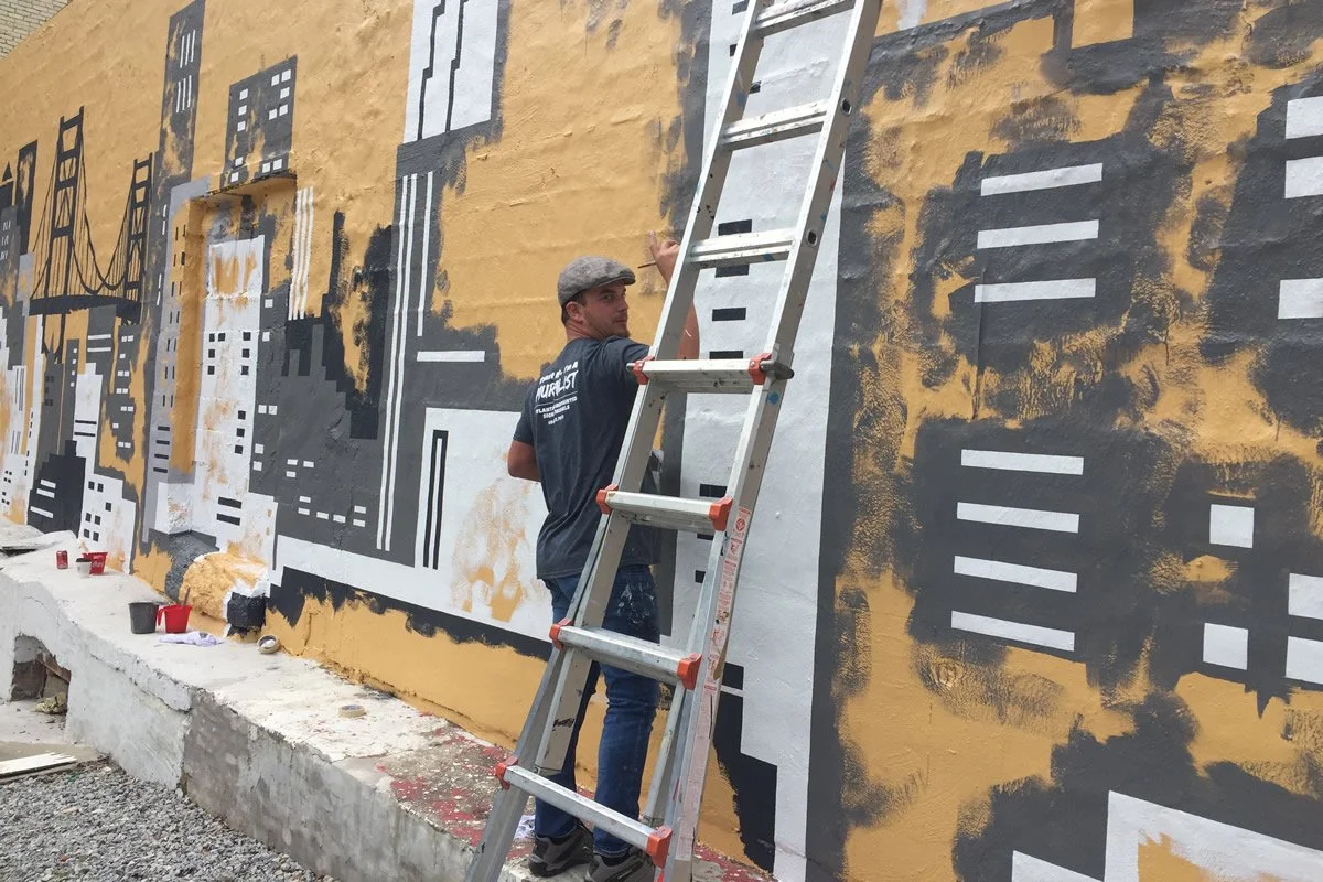 A man standing on a ladder painting a mural of a city skyline on a yellow wall. The mural depicts buildings and bridges in black, white, and gray. Paint cans are on the ground, some opened.