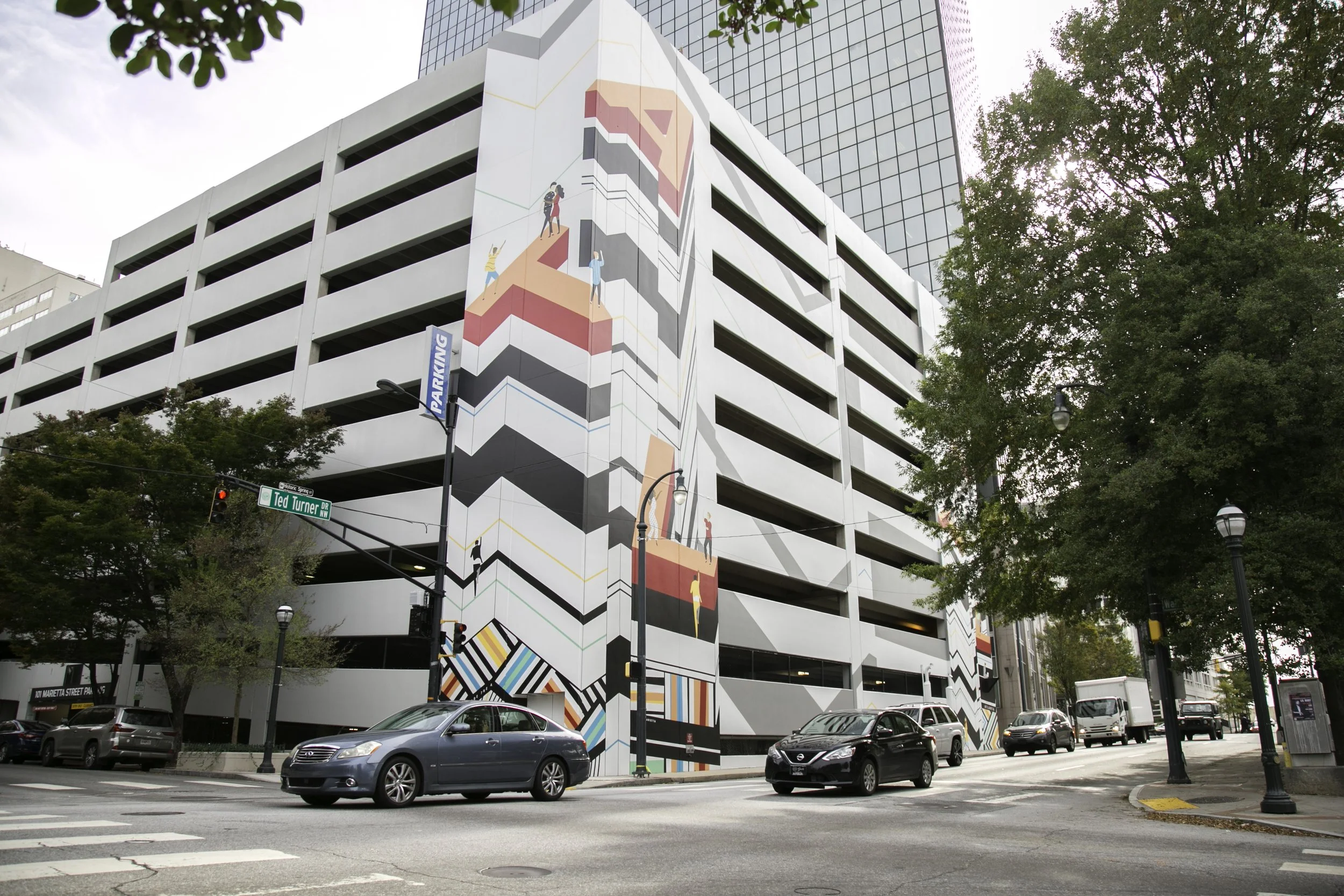 Multi-level parking garage with a colorful mural depicting people climbing a staircase painted on its exterior wall, surrounded by trees and parked cars at street intersection.