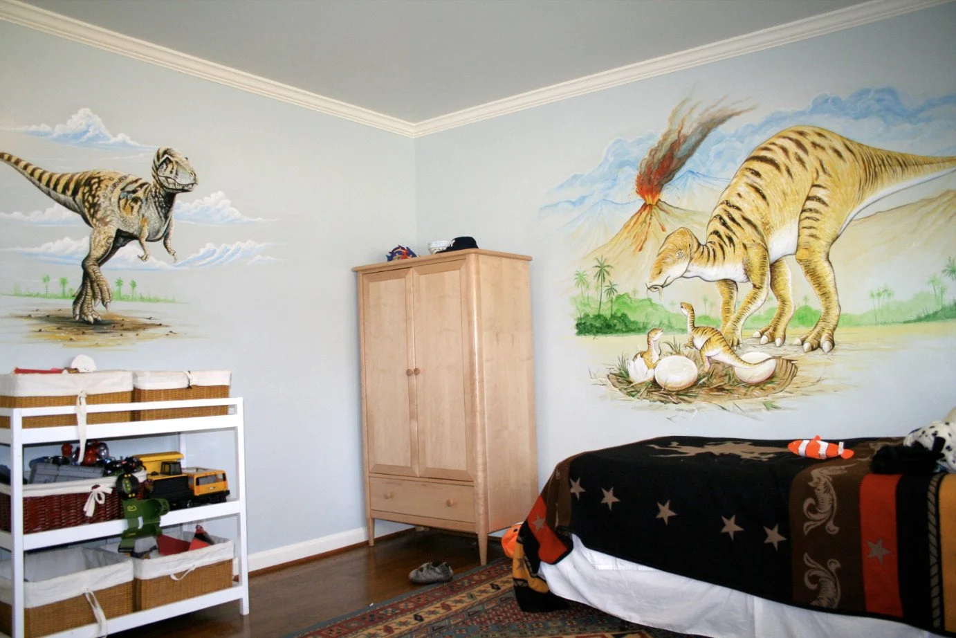 Child's bedroom featuring dinosaur murals on the walls, including a T-Rex on the left wall and a T-Rex with eggs near a volcano on the right wall. There is a white shelf with toy vehicles on the left, a wooden wardrobe in the center, and a bed with a star-patterned blanket on the right.