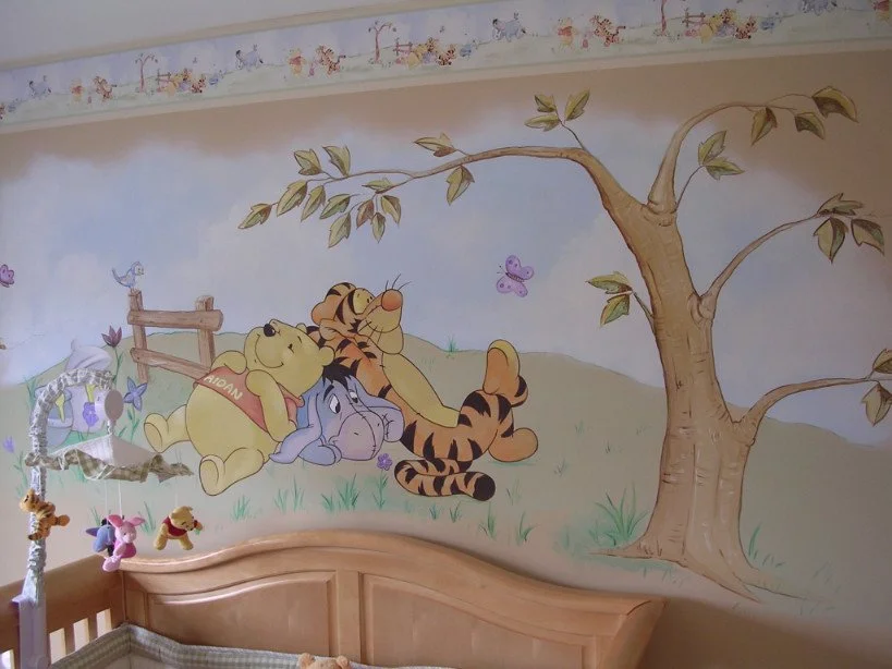 Wall mural featuring Winnie the Pooh, Tigger, and Eeyore resting under a tree, with butterflies and a small fence in the background, in a children's room with stuffed toys on a bed.
