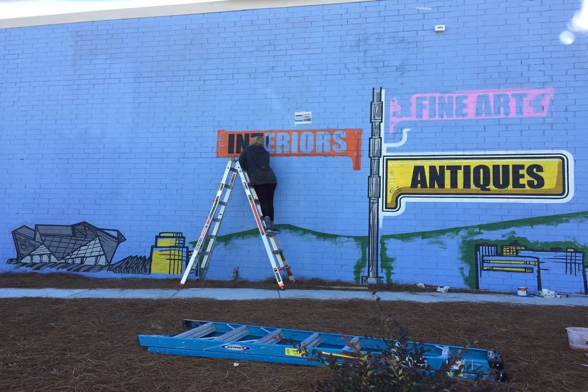A person standing on a ladder painting a mural on a light blue brick wall. The mural features signs for antiques, fine arts, and interior decor, with illustrations of furniture and landscape elements.