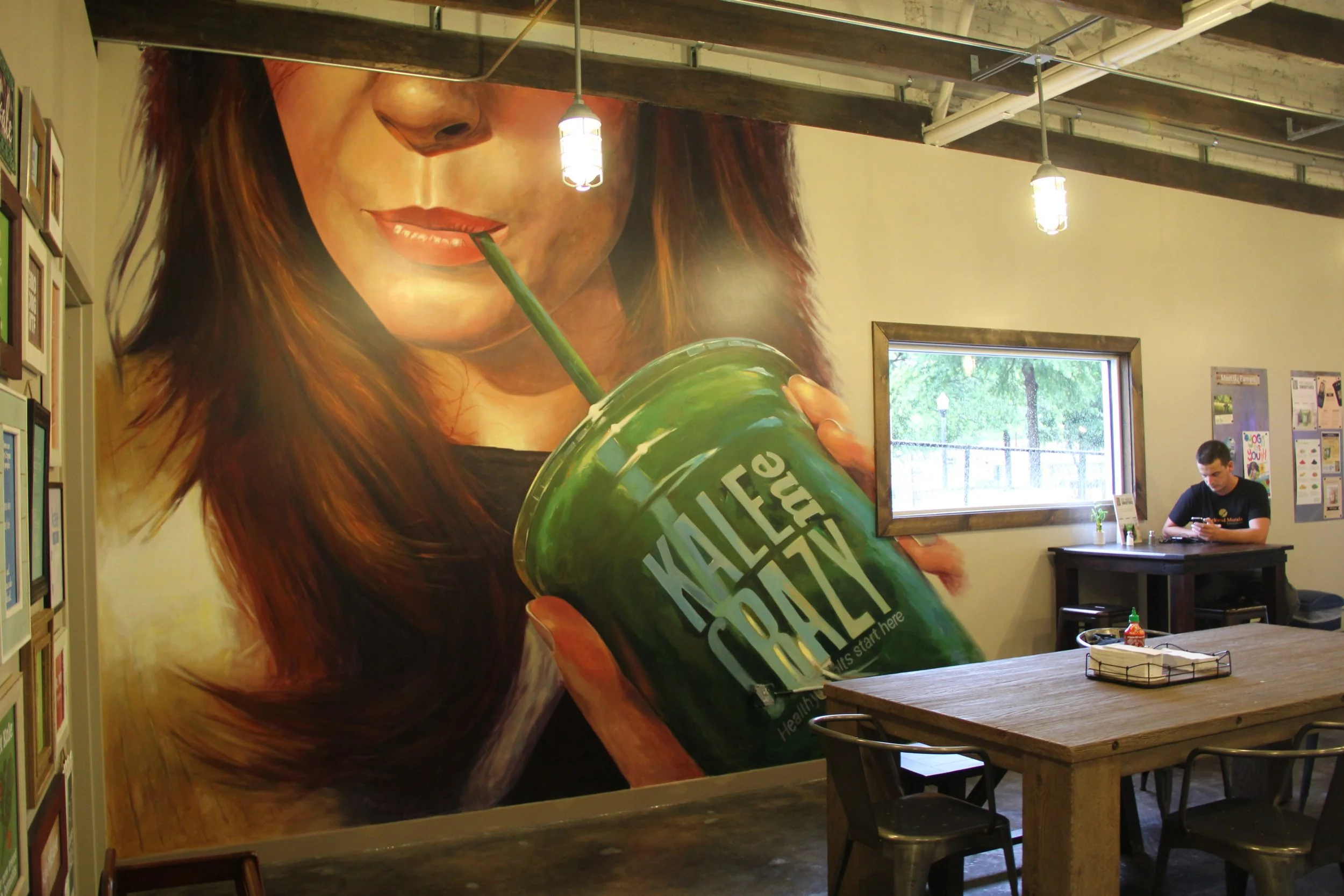 Large mural of a woman with long brown hair sipping from a green beverage cup with a straw, inside a cafe with wooden tables and a window showing trees outside.