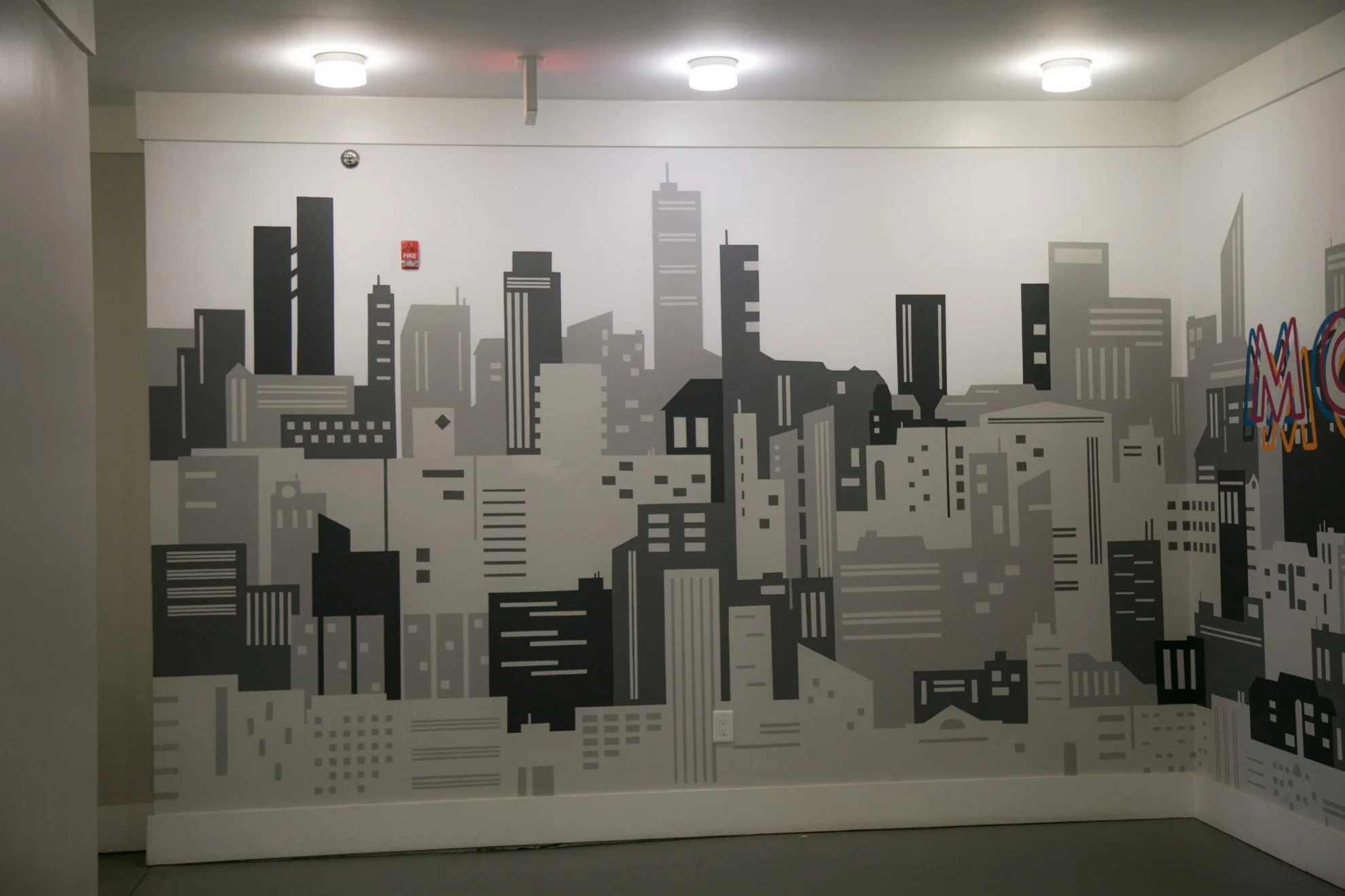 Wall mural of a city skyline in gray tones in an indoor space. There are three ceiling lights and a fire alarm on the wall.