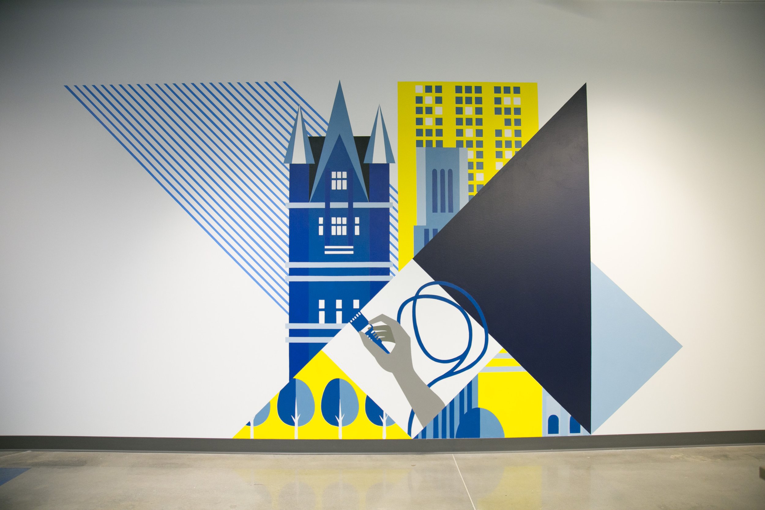A geometric mural featuring stylized buildings and architectural elements in shades of blue, yellow, black, gray, and white, with a hand holding a remote control depicted at the bottom center.