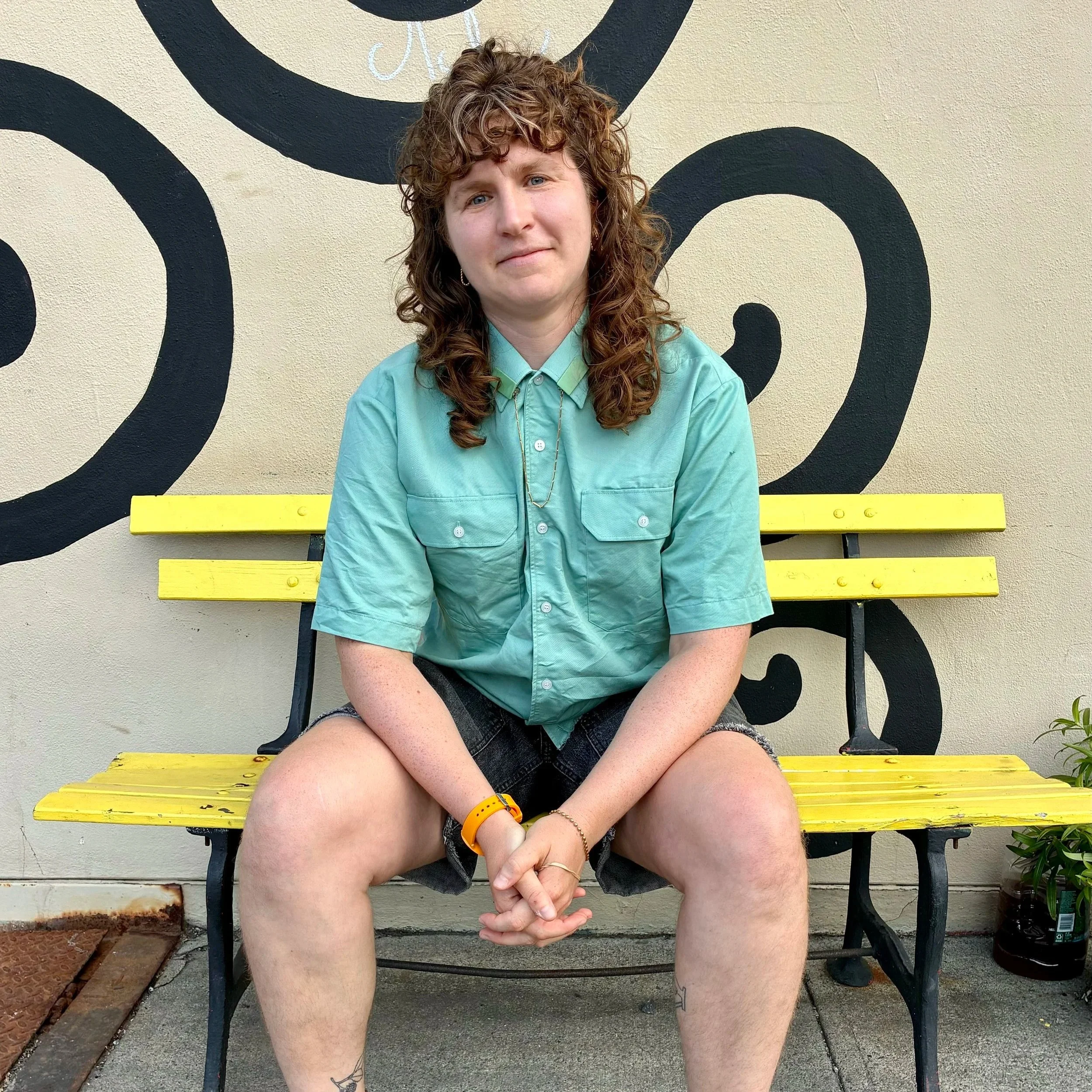 Person with curly hair sitting on a yellow bench in front of a black and white wall mural, wearing a teal shirt and black shorts, with a tattoo on the left ankle and an orange wristband, with a small plant on the right side.