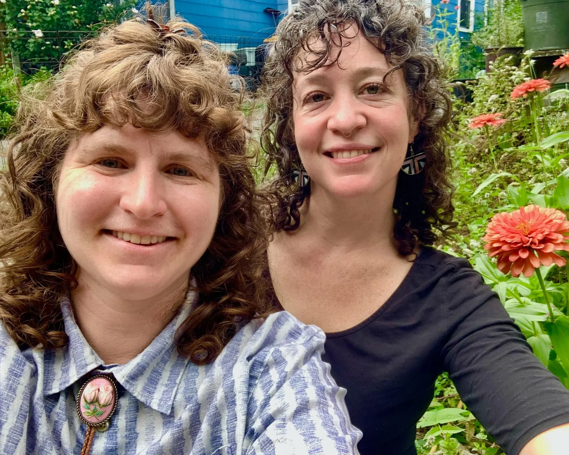 Close-up of two women smiling outdoors in a garden with colorful flowers, one with curly hair and the other with wavy hair.