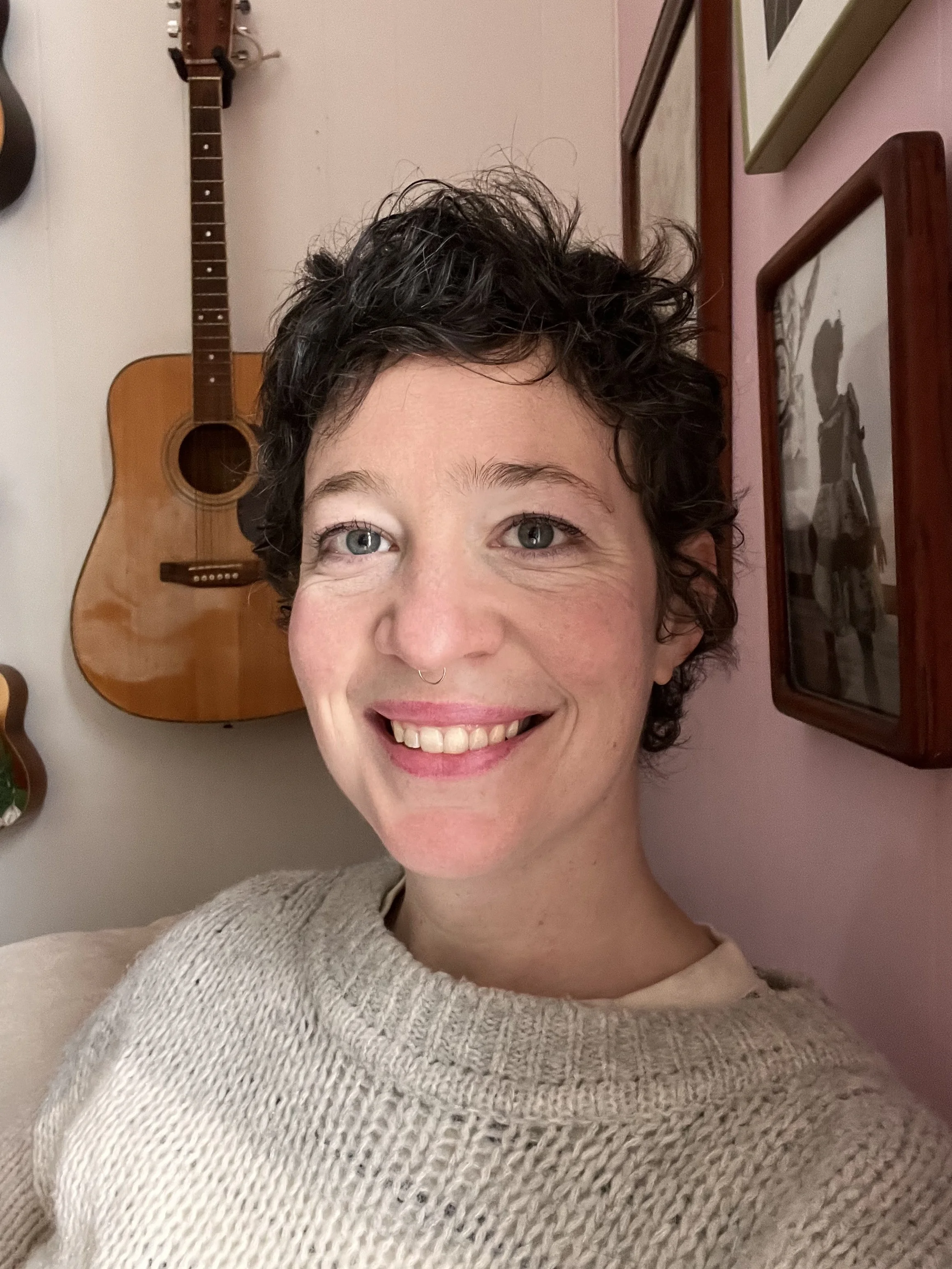 Close-up of a smiling woman with short curly dark hair, wearing a beige knit sweater, standing in front of a wall decorated with framed artwork and a guitar in the background.