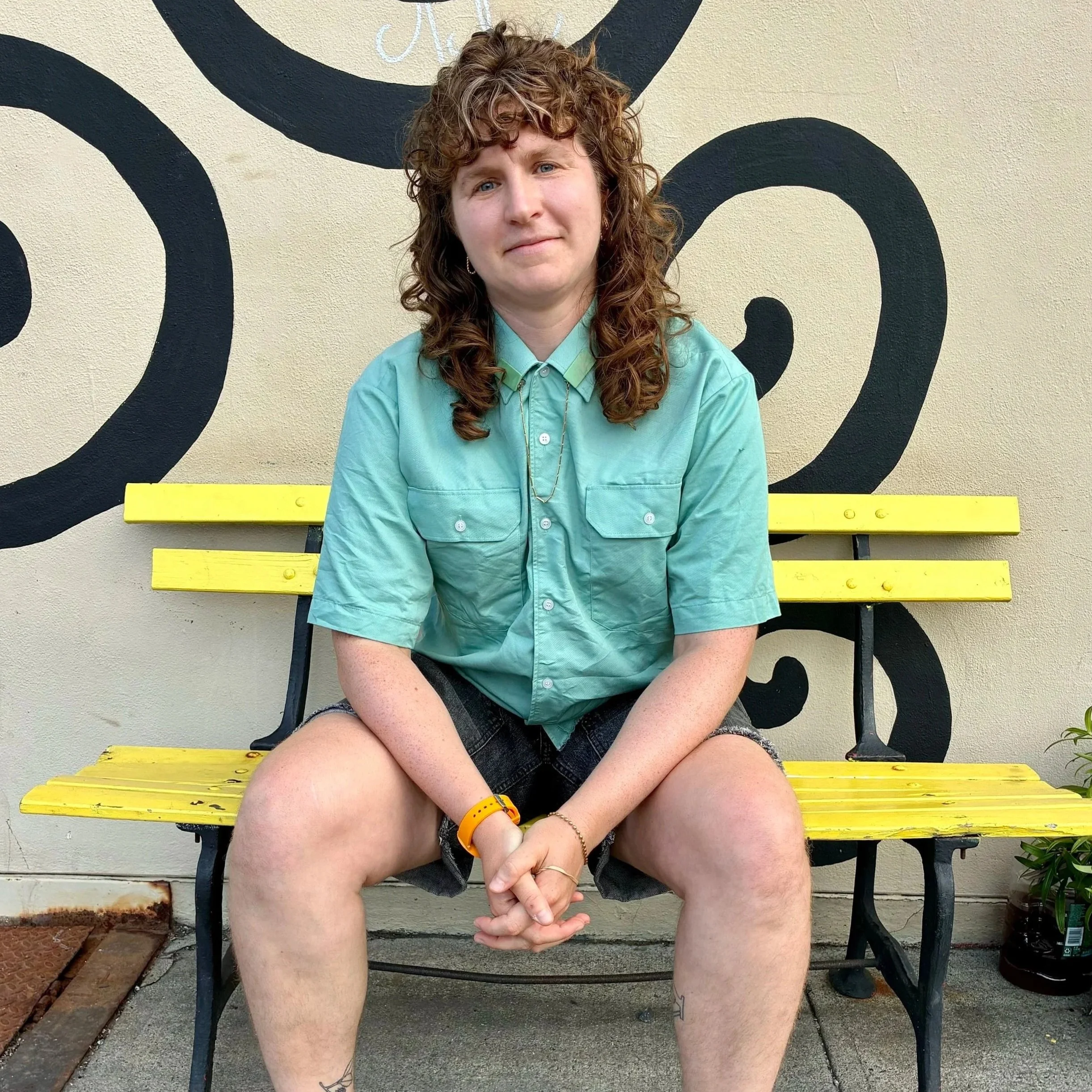 A person with curly red hair sitting on a yellow bench outdoors, wearing a teal shirt, black shorts, and yellow wristbands, with a mural painted on a beige wall behind them.