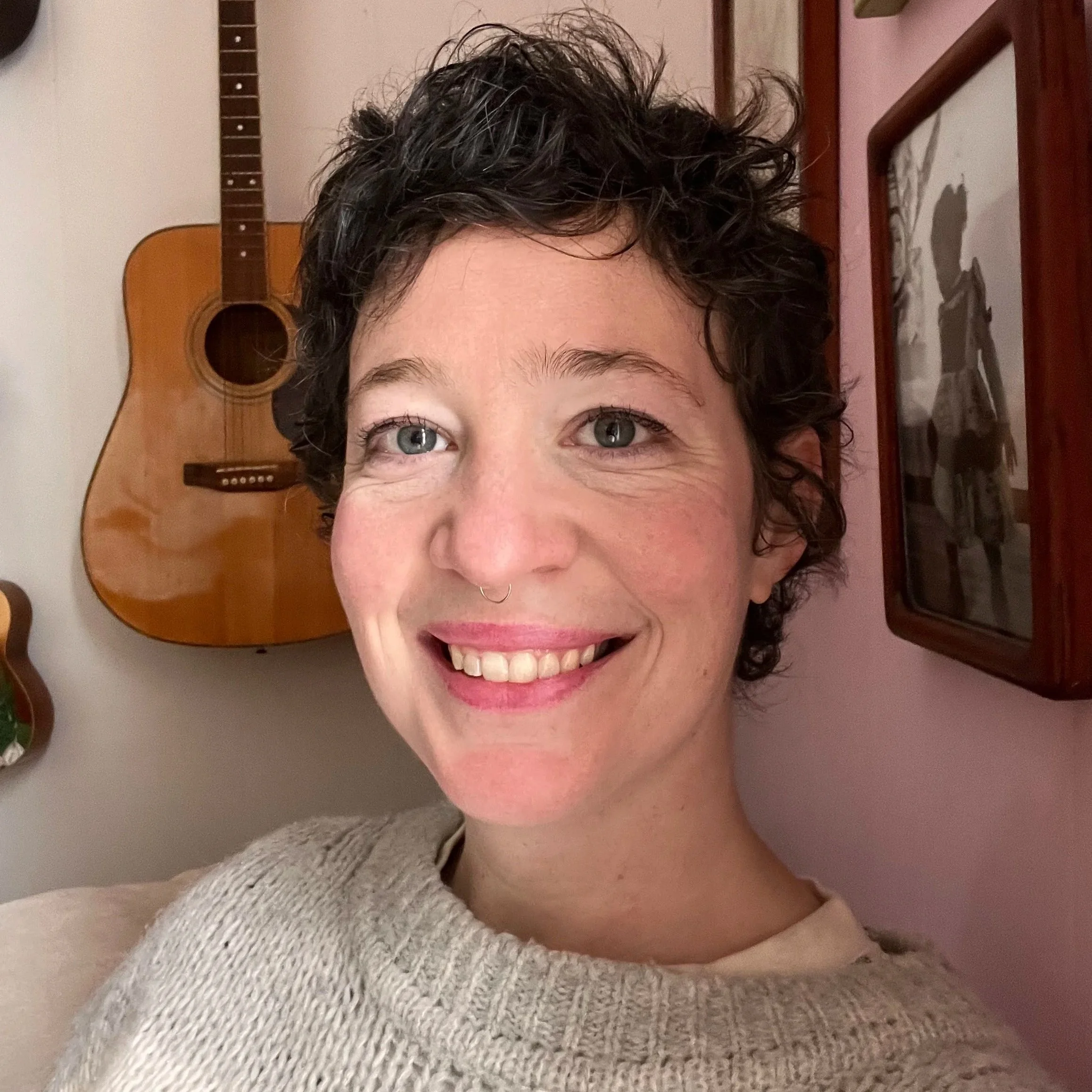 Close-up of a smiling woman's face, showing her eyes, nose, and mouth, with a guitar hanging on the wall in the background.