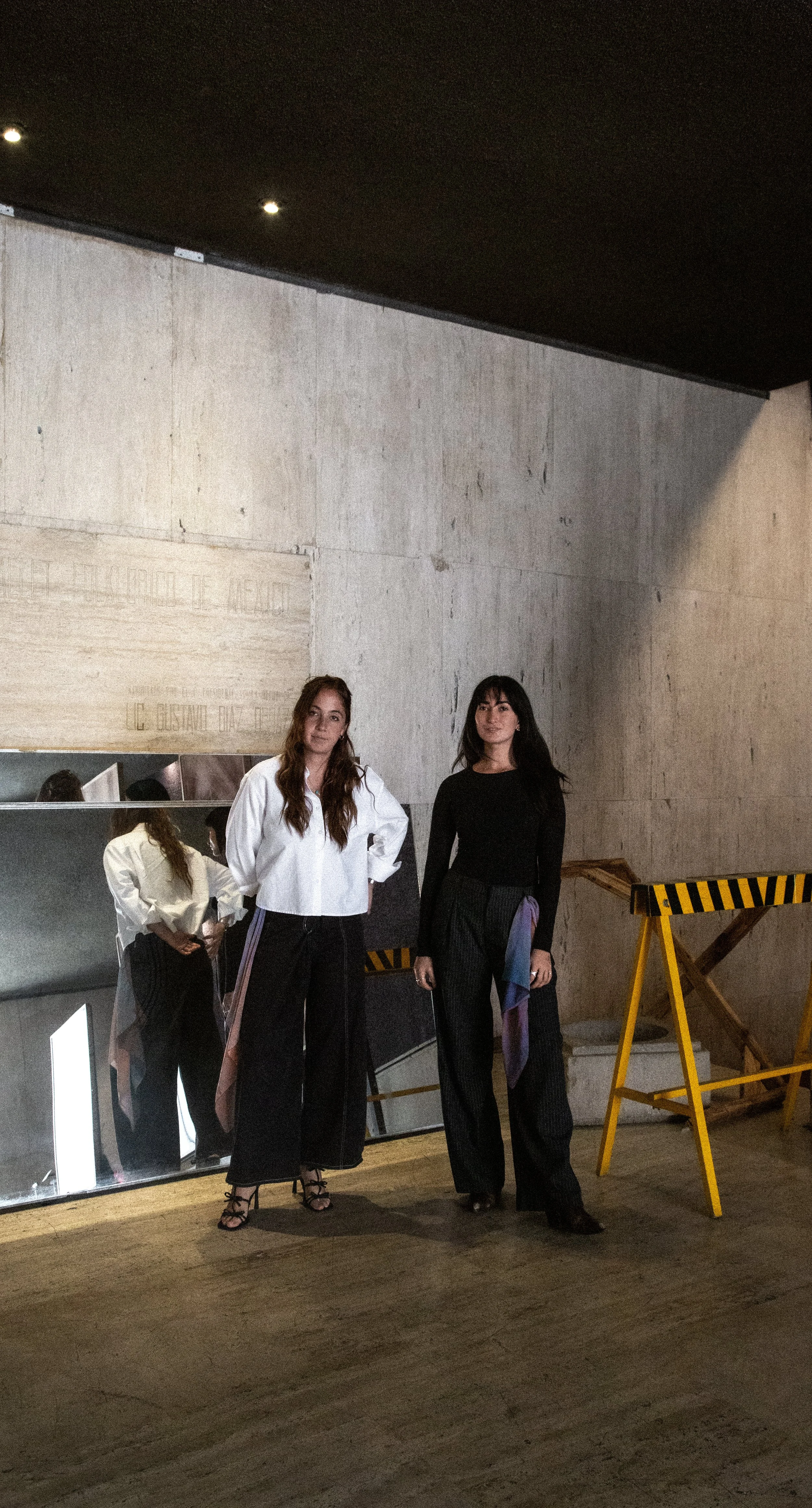 Dos mujeres jóvenes posando en un espacio interior con paredes de concreto y un espejo que refleja parte de ellas. Una de ellas lleva blusa blanca y pantalones negros con gafas y tacones, la otra lleva blusa negra y pantalones con un pañuelo de colores. Hay una estructura amarilla y negra en el fondo.