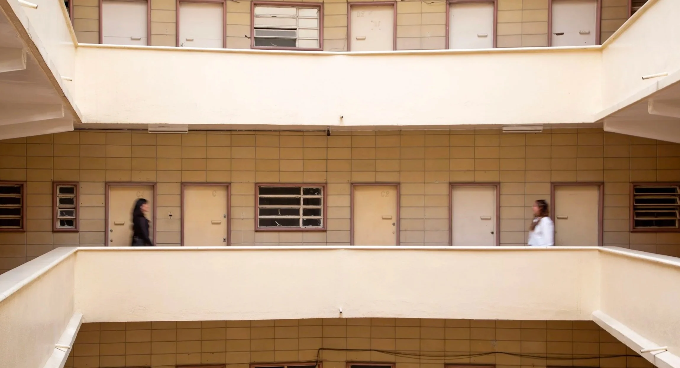 Vista de dos personas caminando en un pasillo de un edificio de apartamentos de varios pisos y paredes de azulejo amarillo, con ventanas y puertas en cada niveles.