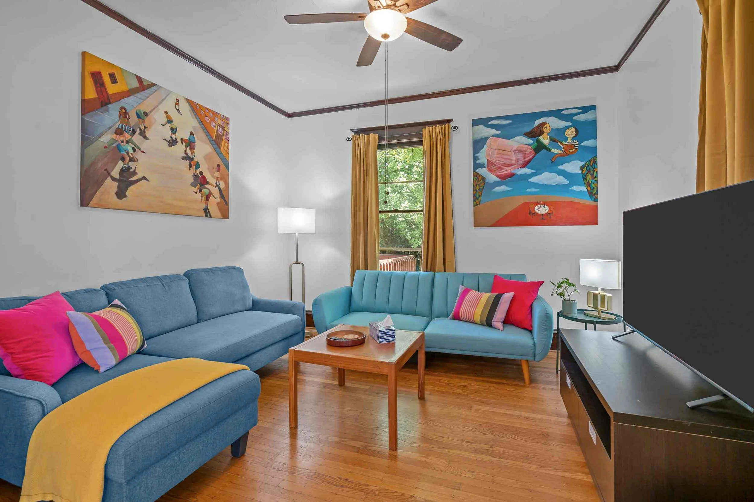 Living room with blue and teal sofas adorned with colorful pillows, wooden coffee table, yellow and striped throw blankets, and wall art featuring children at play and a girl floating with a guitar on a cloud, with yellow curtains and a window showin