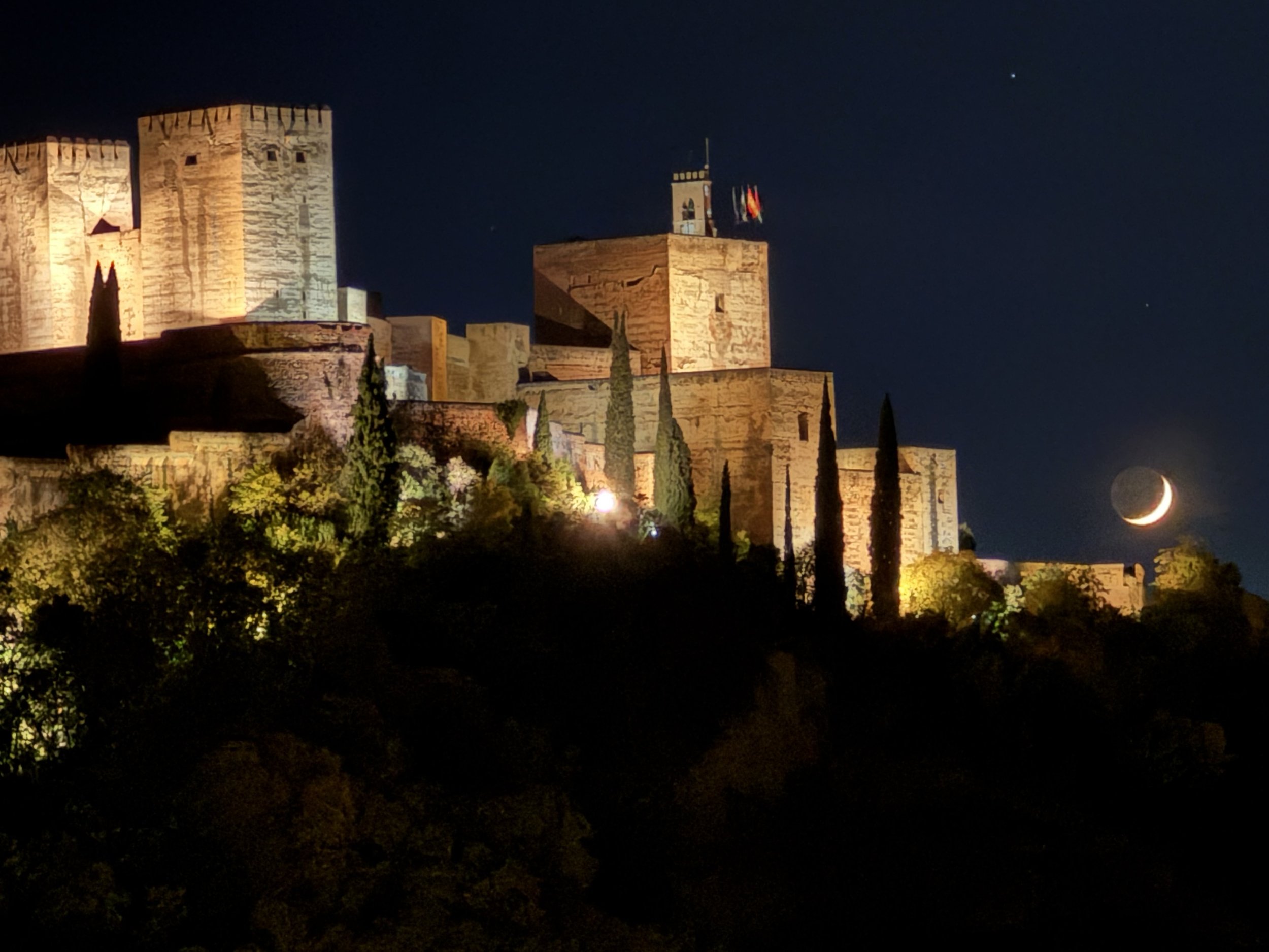 Nighttime view of a historic castle illuminated with warm lights, with a crescent moon in the dark sky to the right.