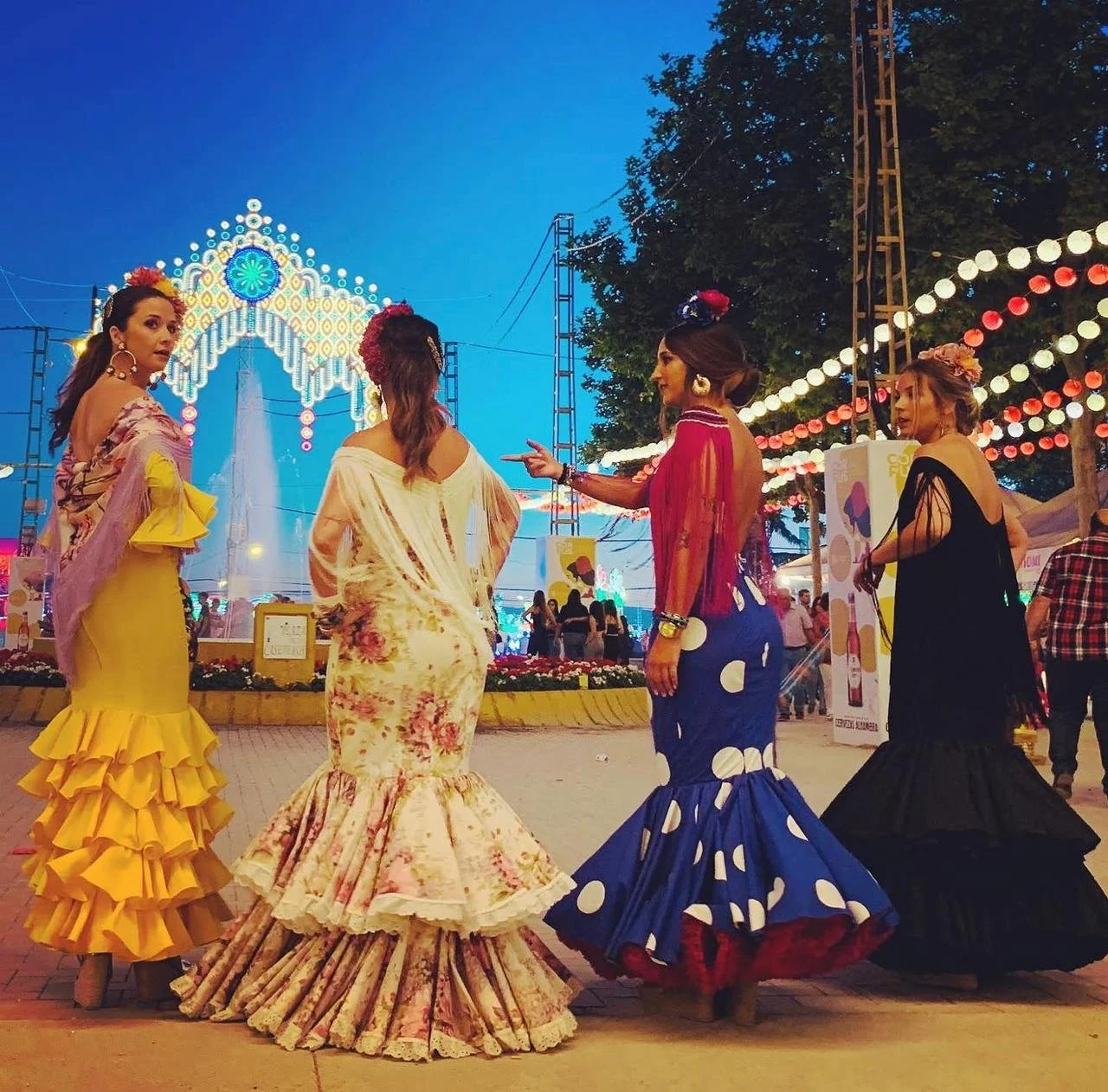 Four women wearing colorful traditional flamenco dresses with ruffles, dancing at an outdoor festival during twilight. The background shows festive lights, a decorated arch, and a crowd.