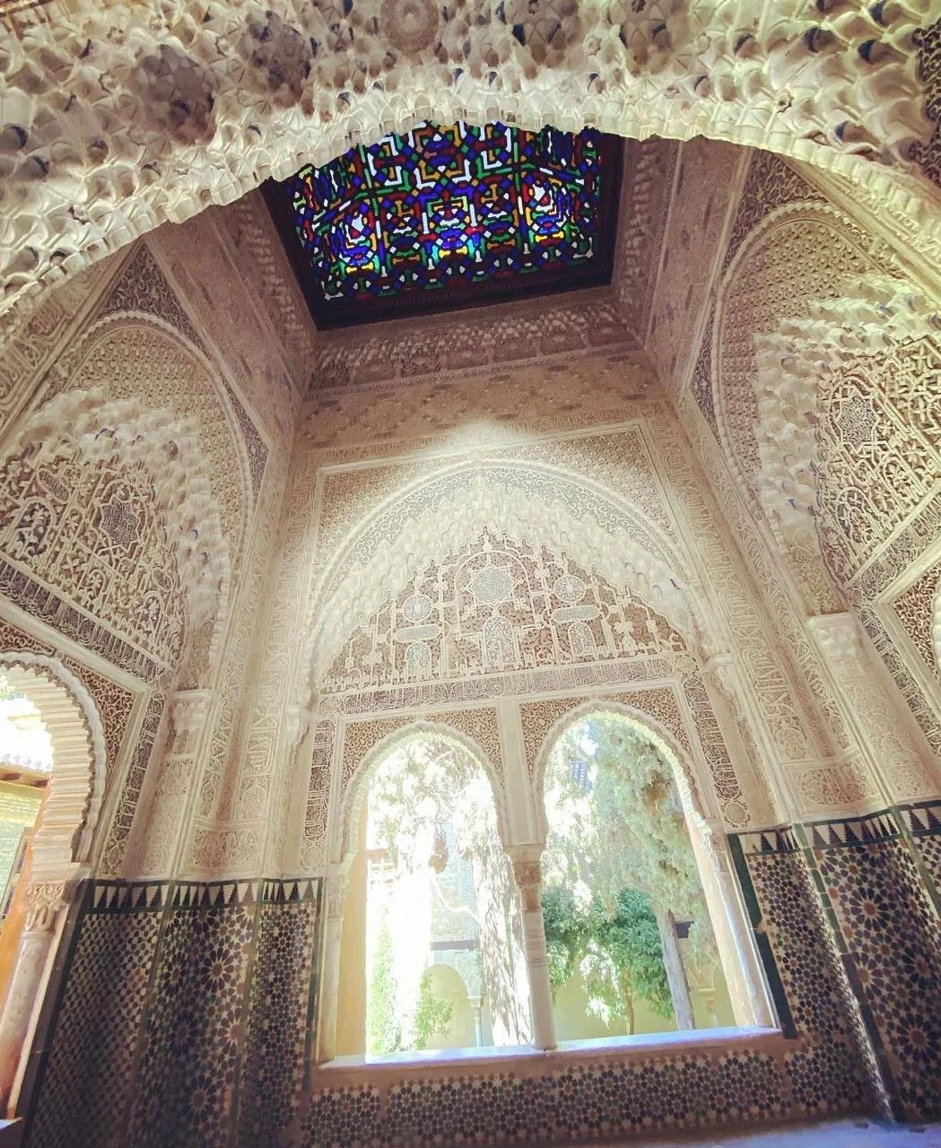 Interior view of a room with ornate Islamic architecture, featuring detailed carvings and calligraphy on the walls, a stained glass ceiling with vibrant colors, and arched windows amounting to a serene, historical ambiance.