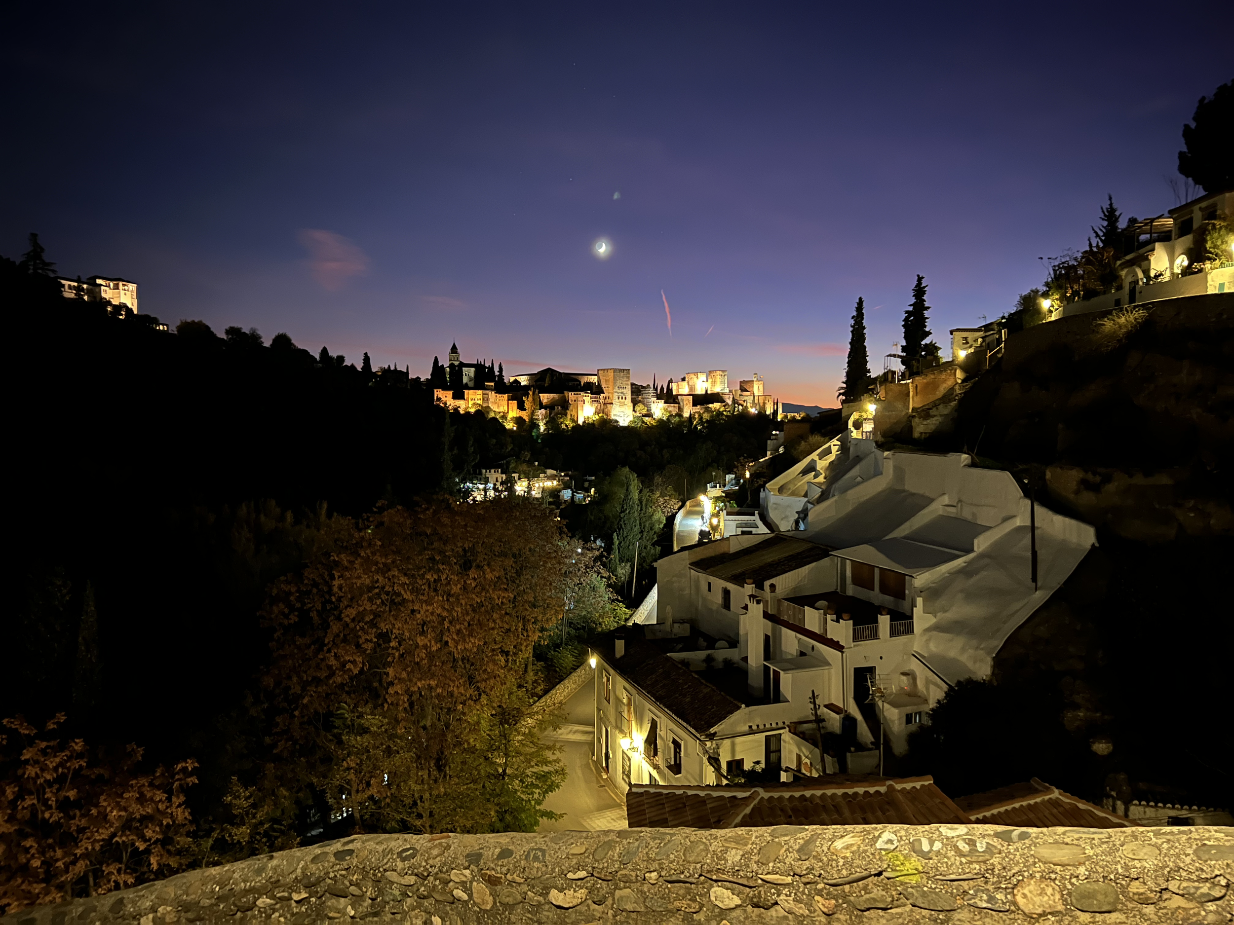 Nighttime view of a hilly cityscape with illuminated historic buildings, modern houses on the hillside, and a crescent moon with a few visible stars in the sky.