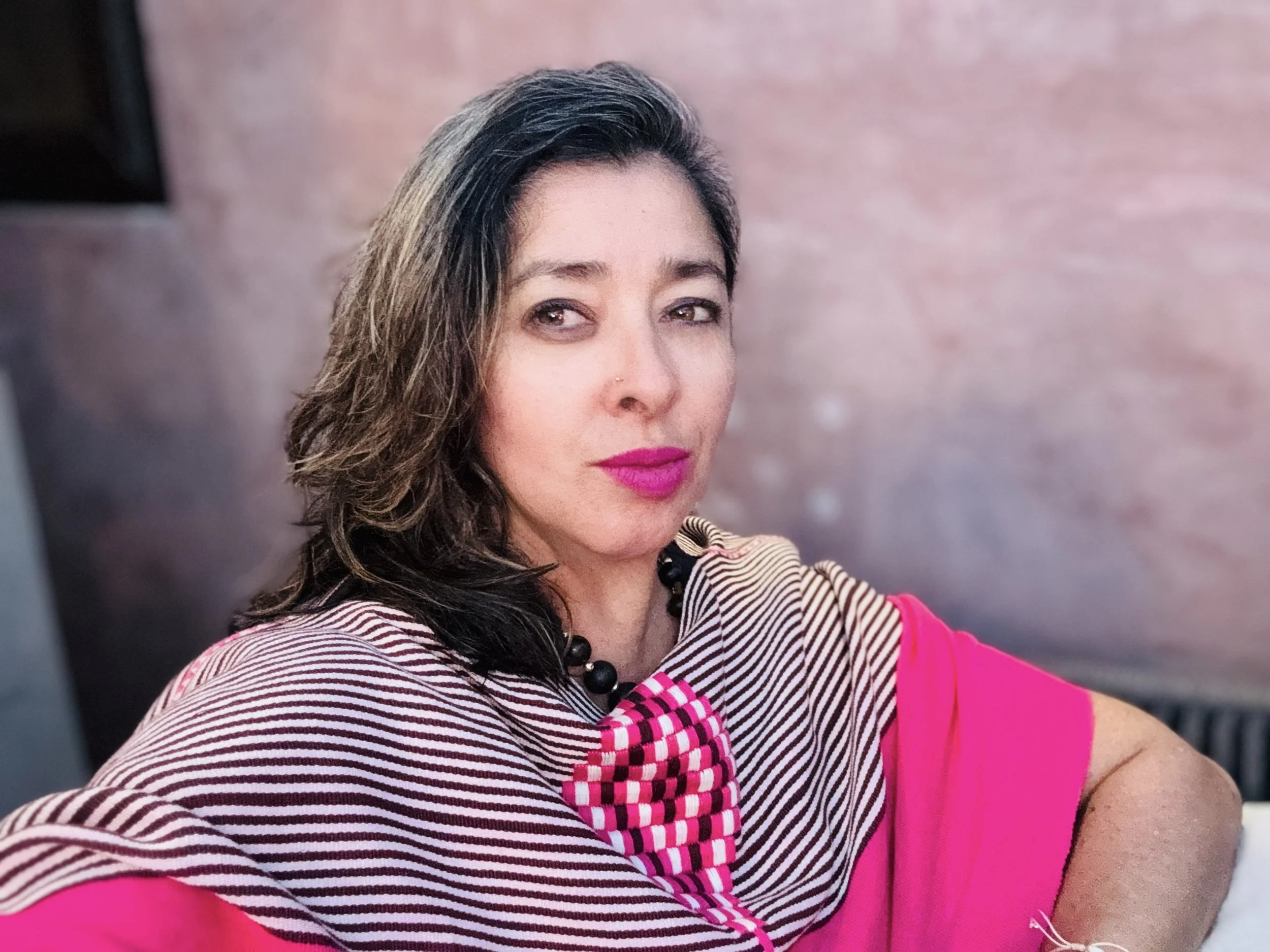Close-up of a woman with brown hair, pink lipstick, and a nose piercing, wearing a striped top and a black beaded necklace, sitting against a pinkish background.