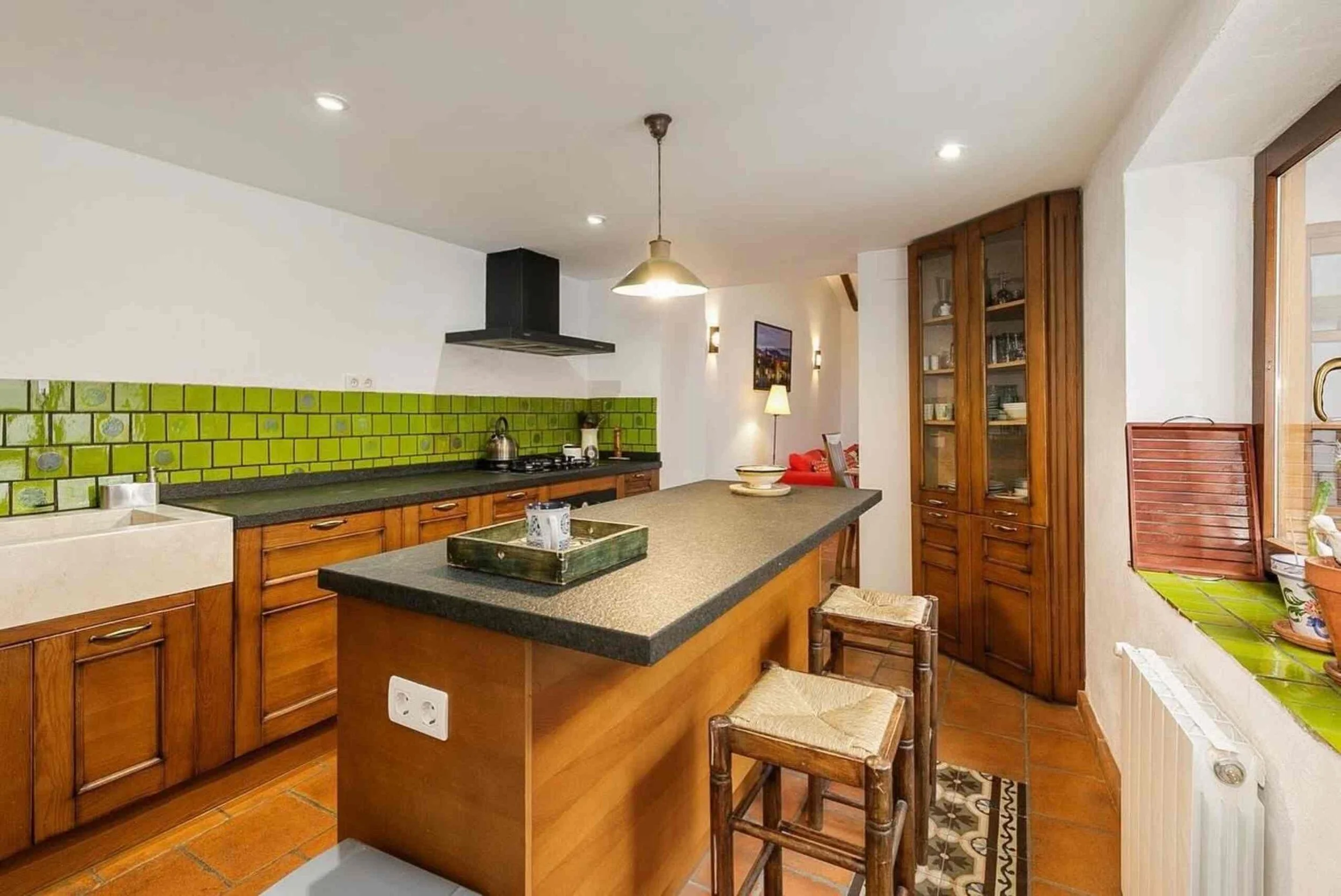 A cozy kitchen with wooden cabinets, a green tile backsplash, a central island with a dark countertop, a stove with a black range hood, and a small dining area with stools. There is a window with green and orange patterned curtains and a door leading
