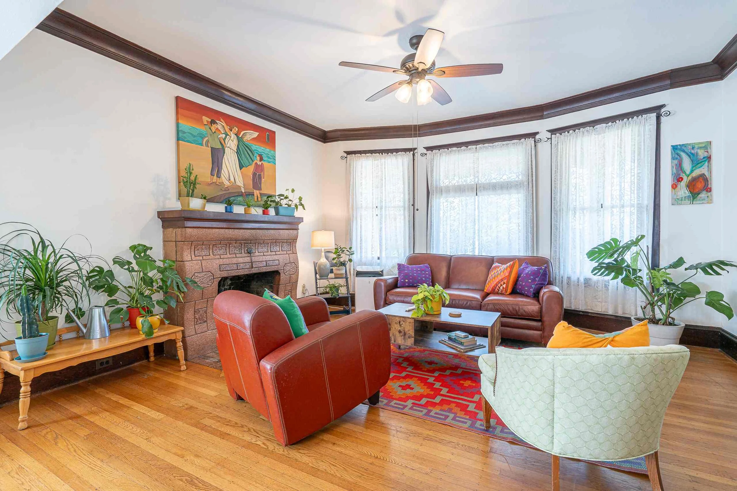 A living room with a brick fireplace, colorful paintings, large windows with white curtains, and plush seating including a red leather armchair, a sofa with colorful pillows, and an off-white armchair, decorated with plants and wooden furniture.