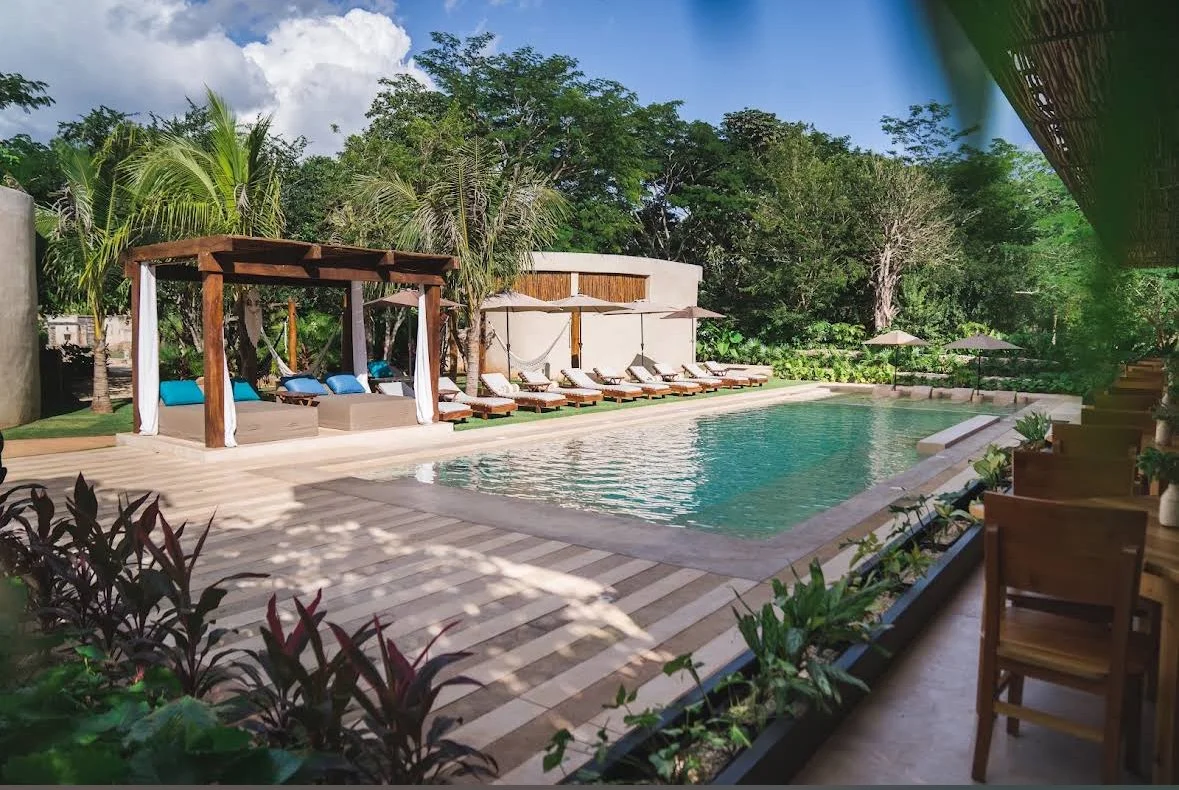 Luxury outdoor swimming pool surrounded by lounge chairs, umbrellas, and lush greenery with tropical plants and trees, under a partly cloudy sky.