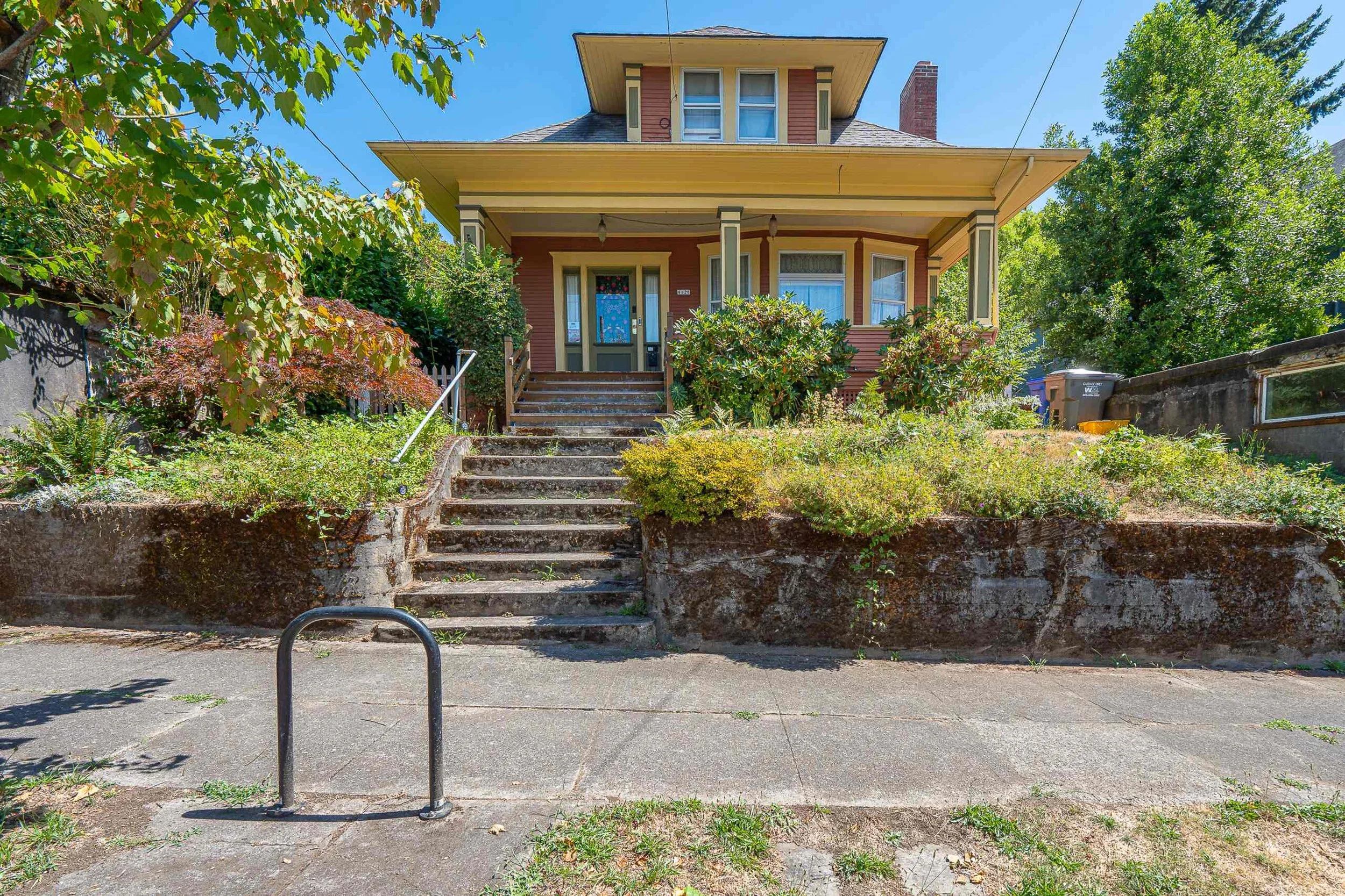 A front view of a two-story house with a porch and steps leading to the entrance, surrounded by trees and greenery, with a pathway and a metal railing in the foreground.