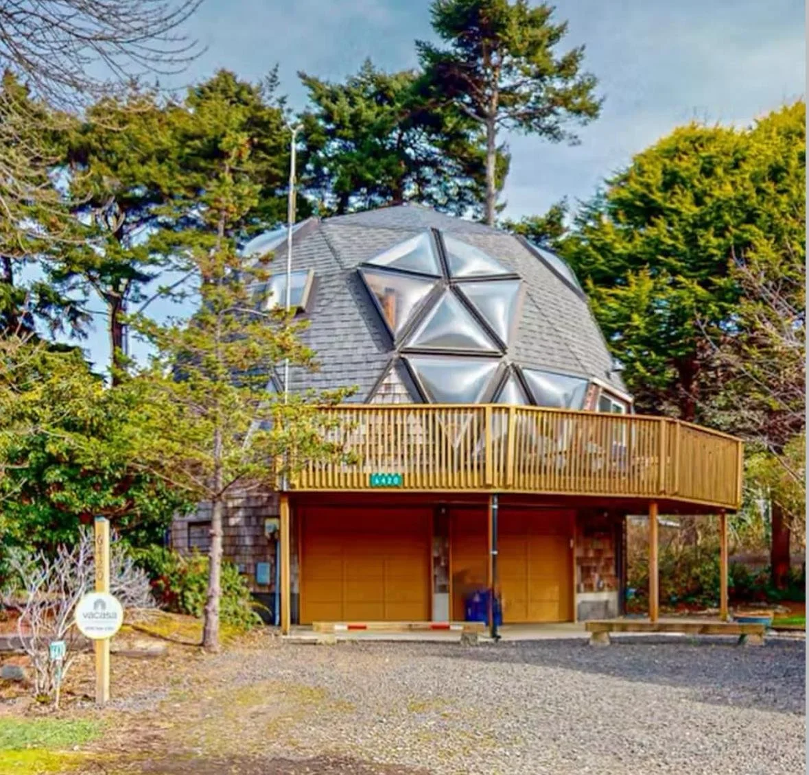 A house with a geodesic dome shaped roof with triangular panels, surrounded by trees and a wooden balcony in the front.