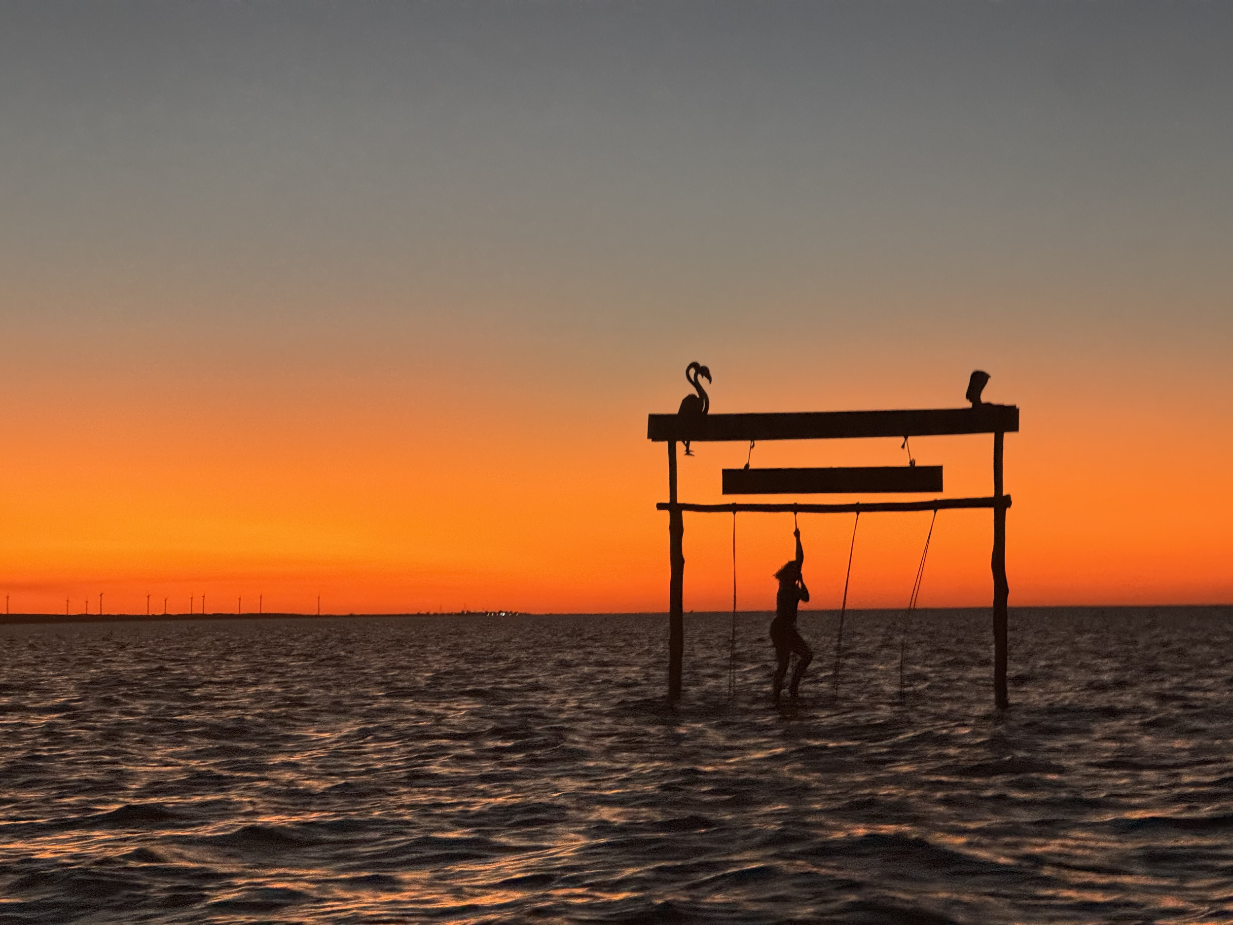 Silhouette of a person sitting on a swing set near water during sunset, with a backdrop of a colorful sky with shades of orange, yellow, and gray.