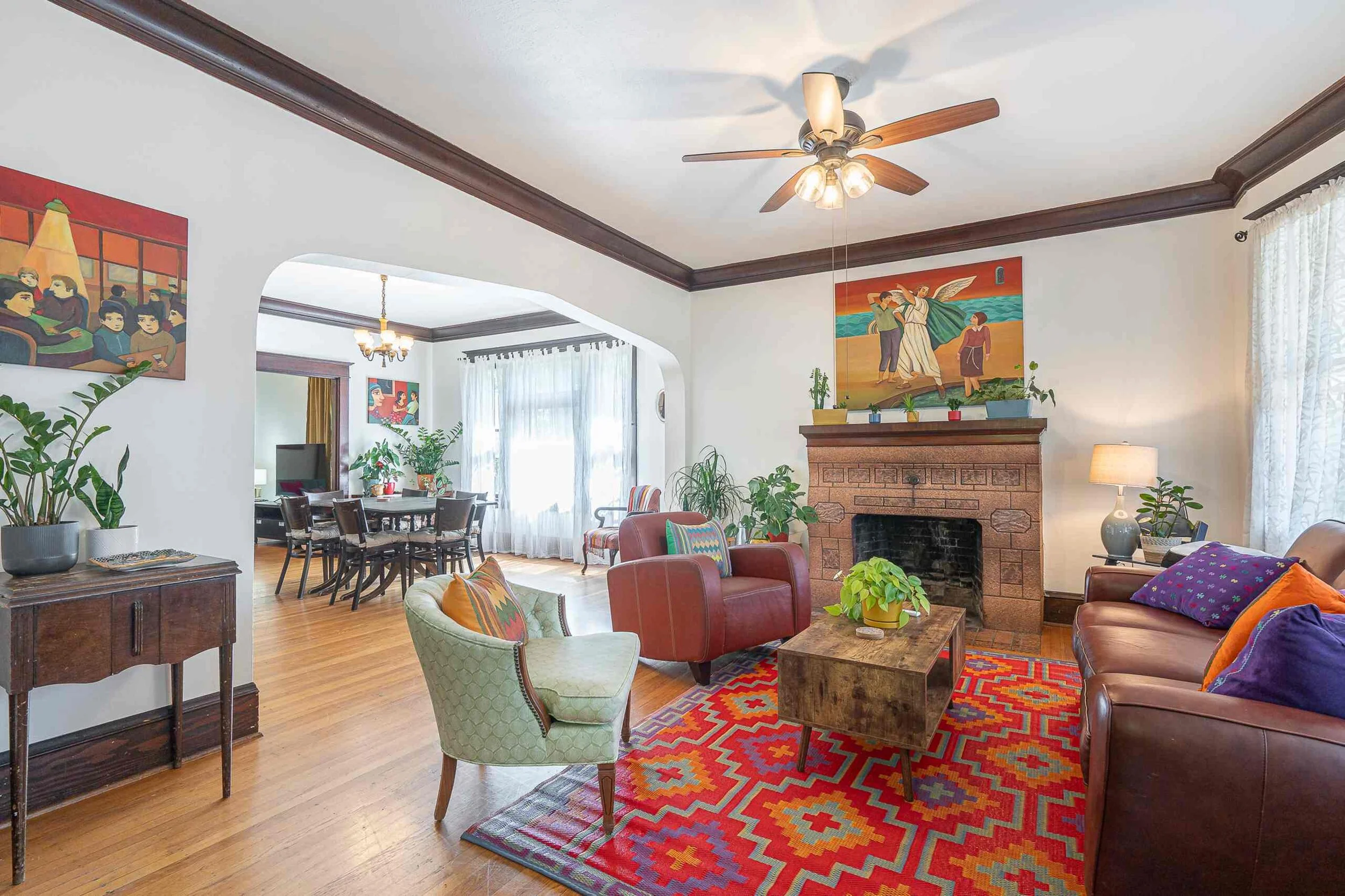 A cozy living room with colorful furniture, plants, wall art, and a brick fireplace.