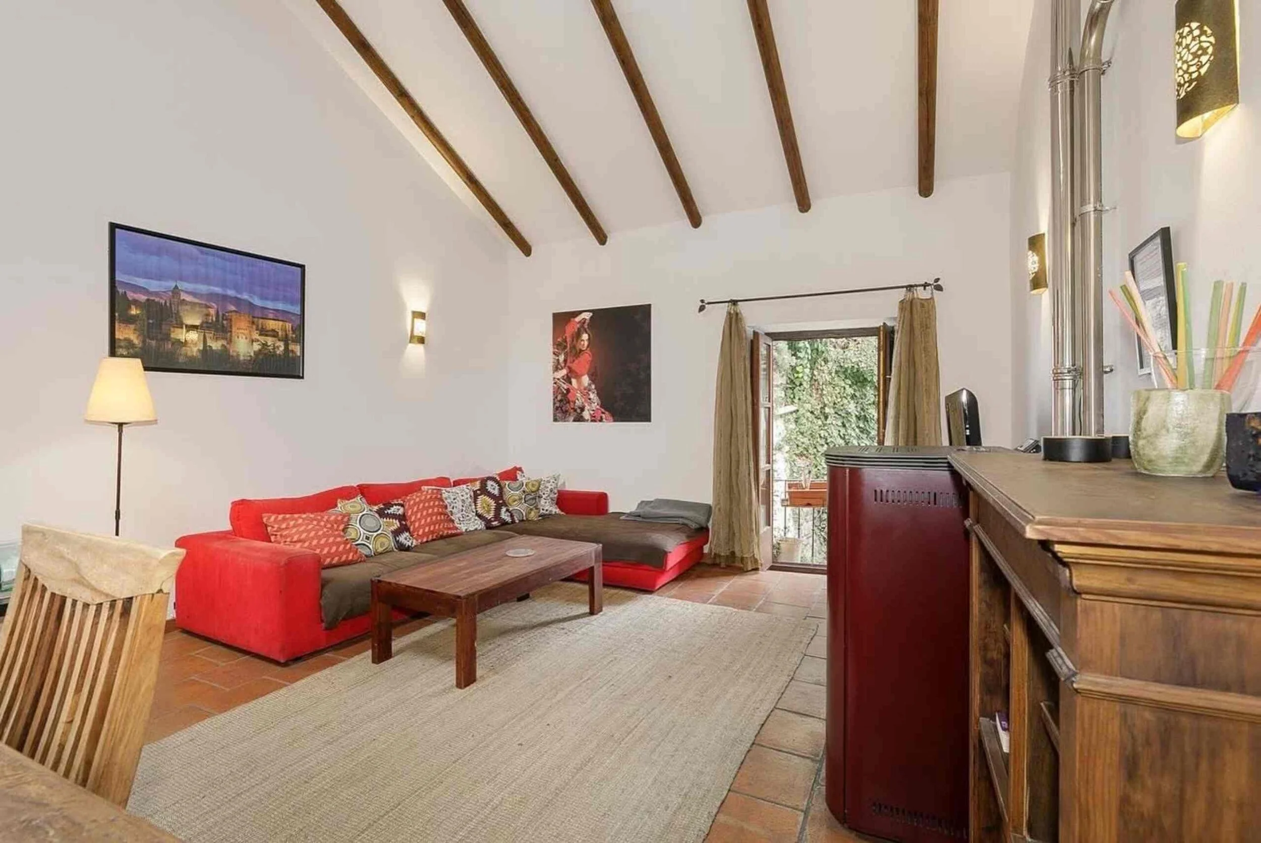 Living room with a red sectional sofa with patterned pillows, wooden coffee table, beige rug, wooden beams on ceiling, wall decoration including a framed picture of a cityscape, a portrait of a woman, a lamp, and a window with curtains showing greene